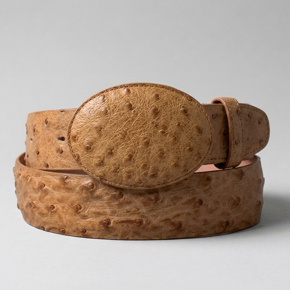 A Los Altos amber tan ostrich leather belt  with a matching oval buckle, coiled on a light gray background.
