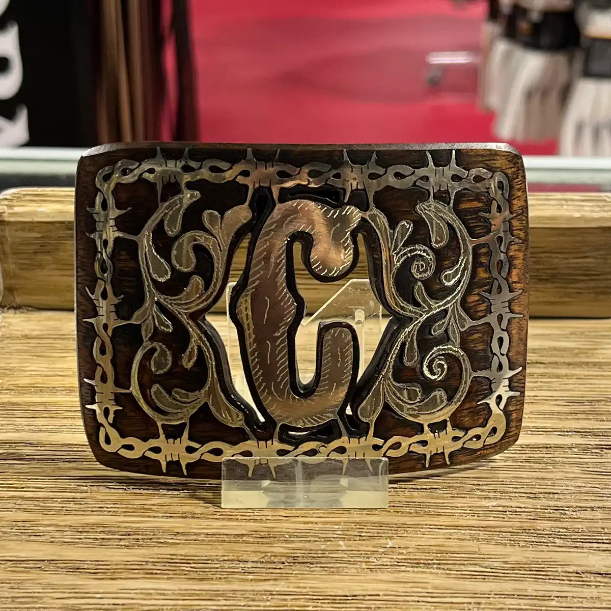 A rectangular wooden belt buckle with an engraved Celtic-style scroll pattern and a central raised letter 'G', displayed on a clear stand on a wooden surface.