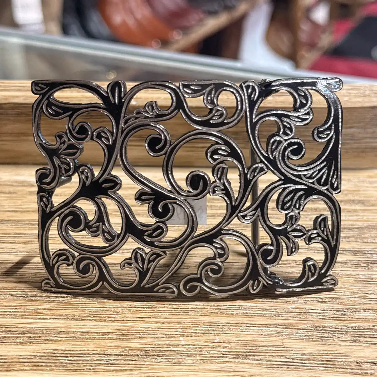 In-store counter display view of the 3.5" x 2.5" stainless steel cutout belt buckle showing the deep black background and silver floral filigree.