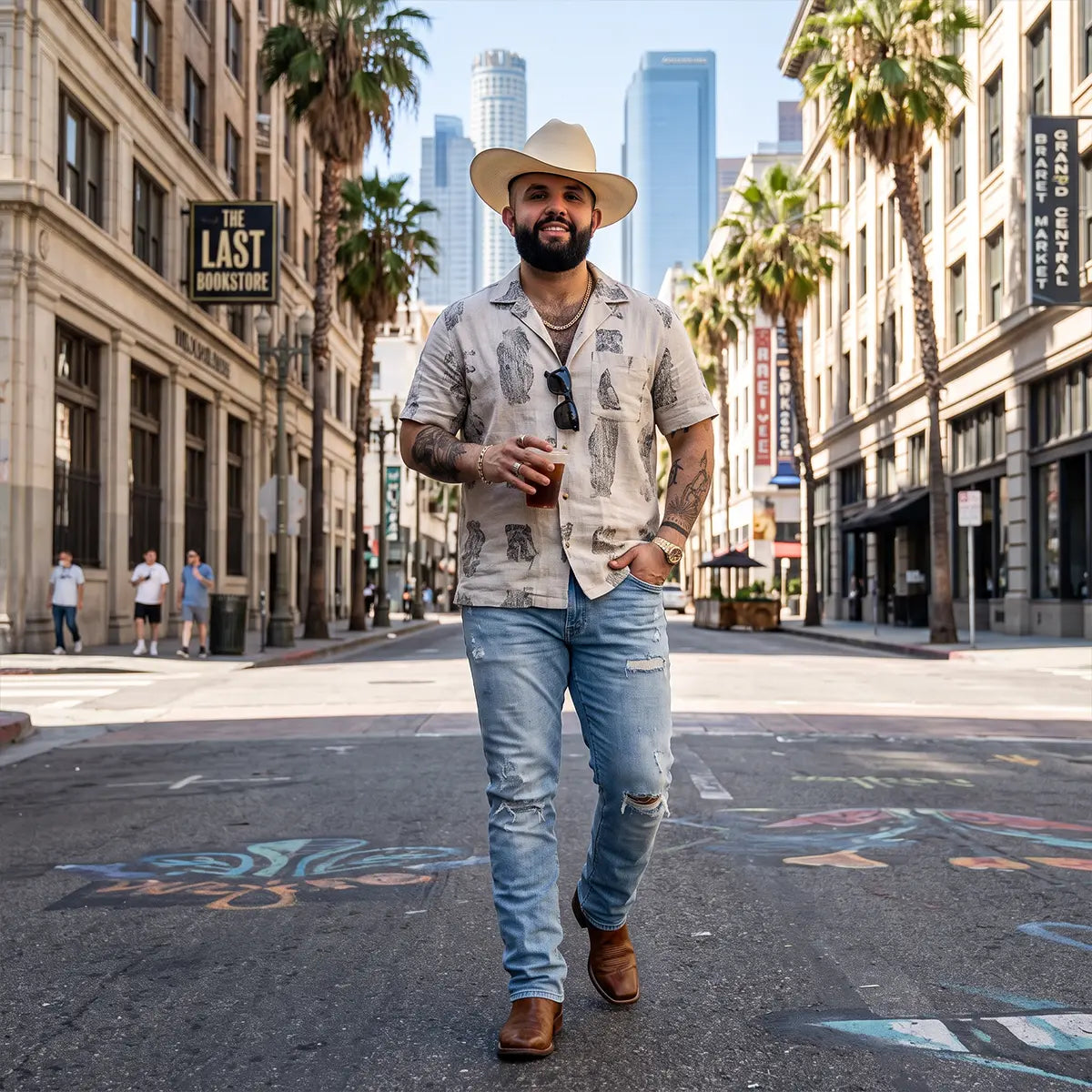 A man wearing a Carin Leon cowboy hat, a light beige patterned short-sleeve button-up shirt, distressed light blue jeans, and brown boots walks down a city street lined with palm trees and buildings.