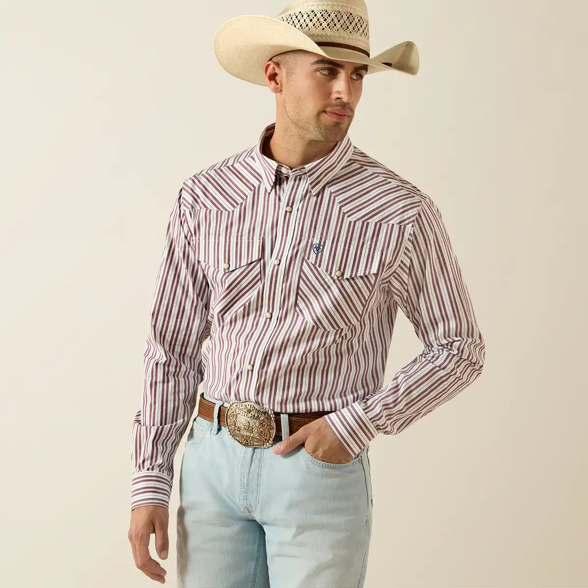 Man wearing a striped Ariat shirt, light blue jeans, and a cowboy hat on a beige background.