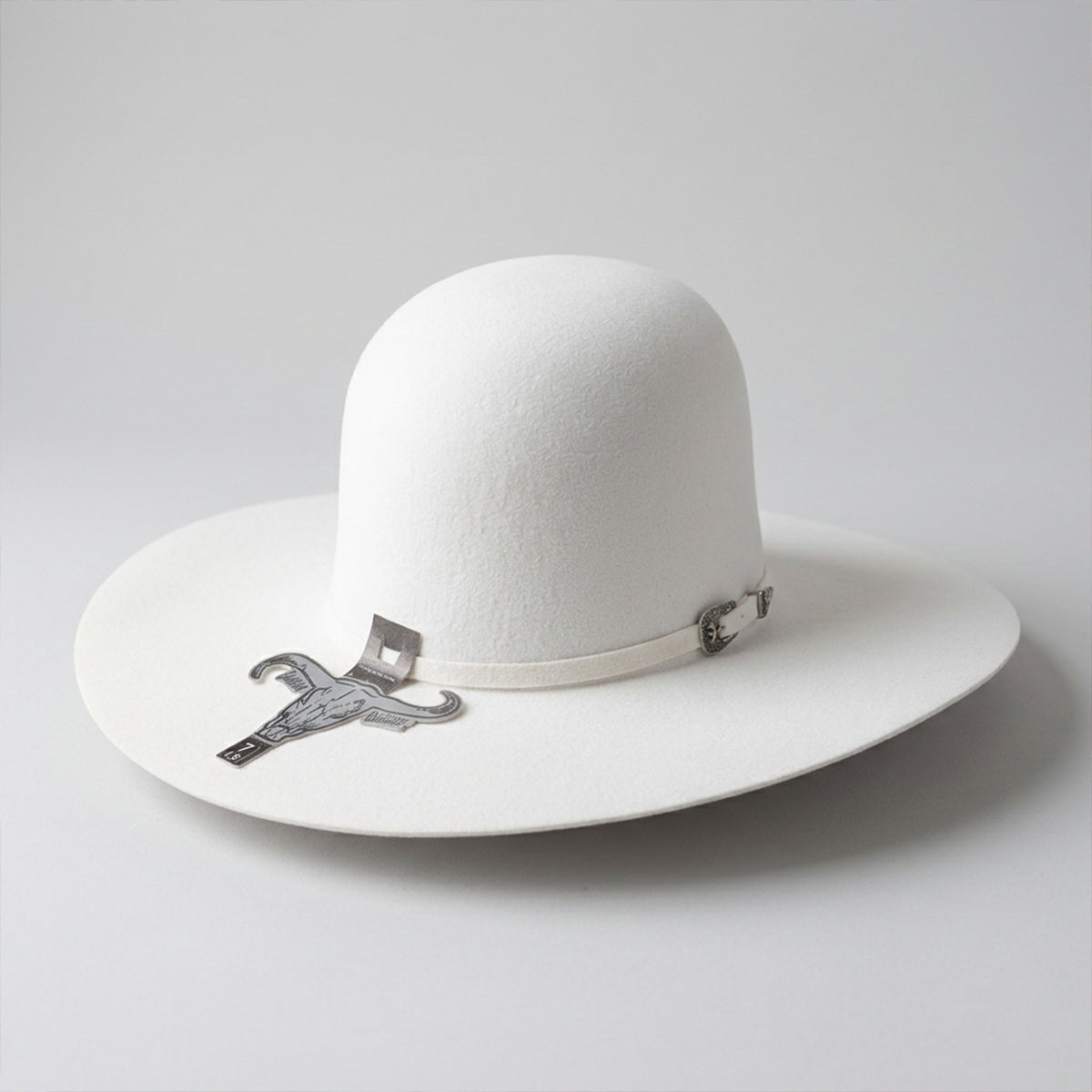 A Cuernos Chuecos white open crown felt western hat with a matching white band and a small silver metal emblem, photographed on a light gray background.