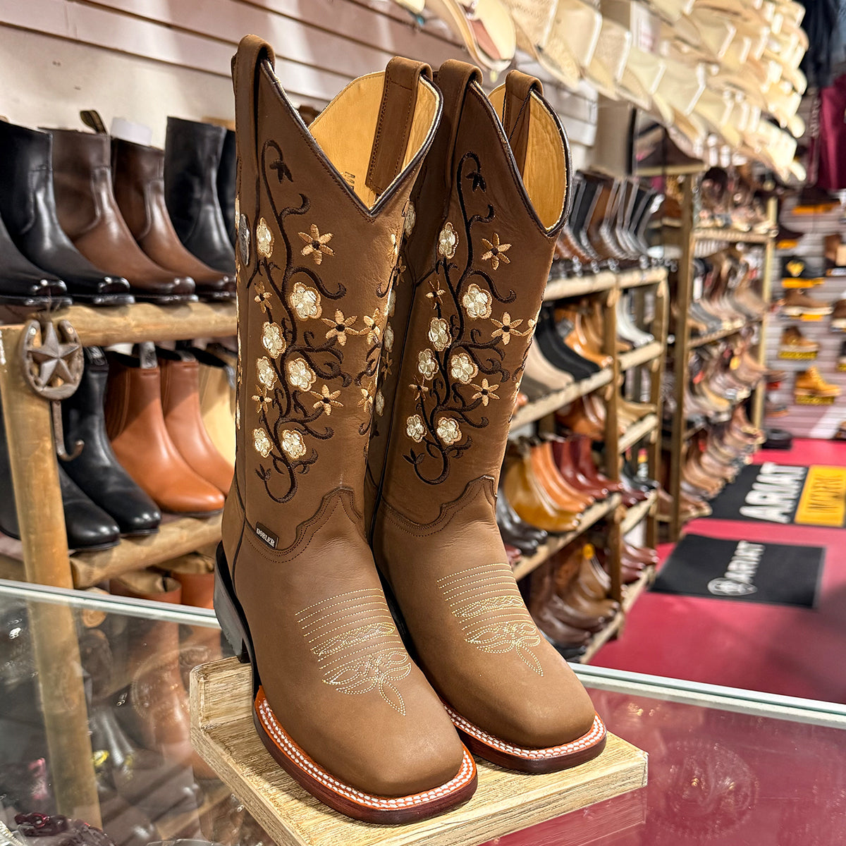 A pair of women's brown leather cowboy boots with floral embroidery, displayed on a wooden stand inside a western store.