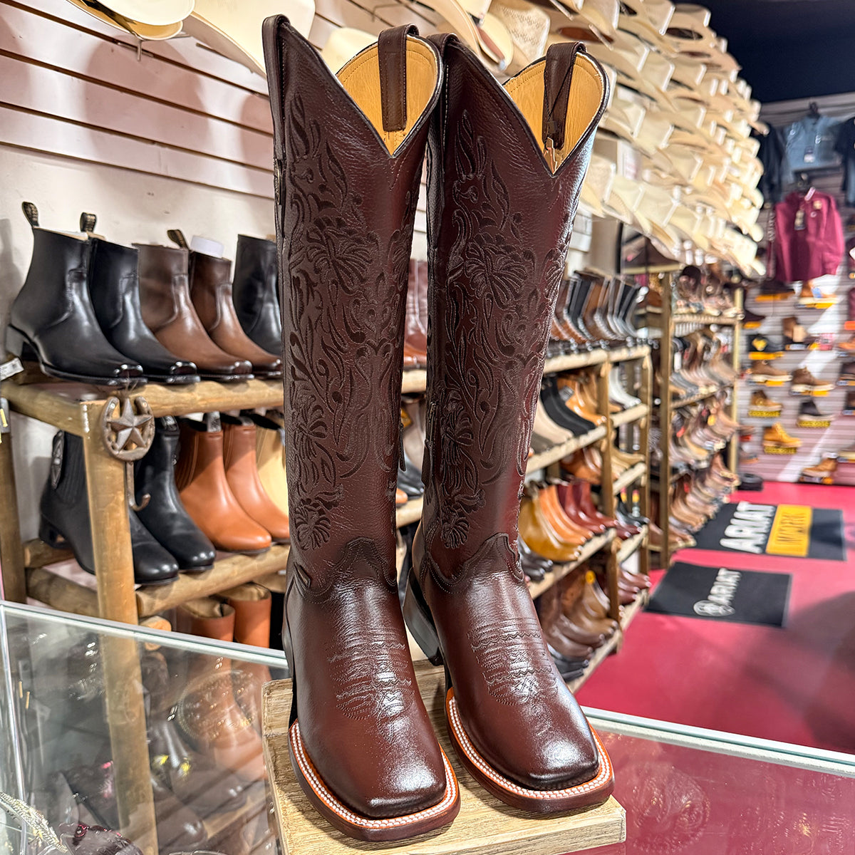 A pair of Doble R women's dark brown tooled leather western square toe boots displayed on a wooden stand inside a western store, with other boots visible on shelves in the background.