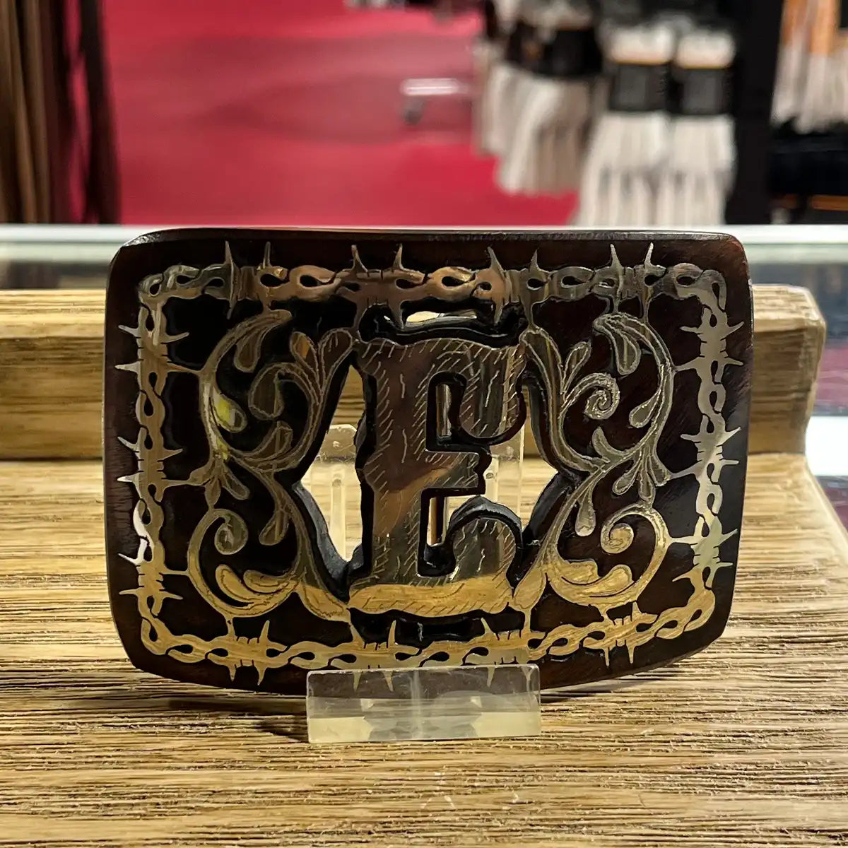 In-store counter display view of the handcrafted mesquite wood belt buckle showing the Monogram E inlay on an acrylic stand.