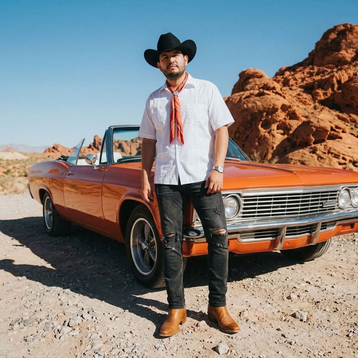 Outdoor lifestyle shot of Regional Mexican singer Eden Muñoz wearing his signature black tejana cowboy hat, short-sleeve white guayabera shirt, red bandana, distressed black jeans, and cognac leather boots in front of a classic orange convertible car in a desert landscape.