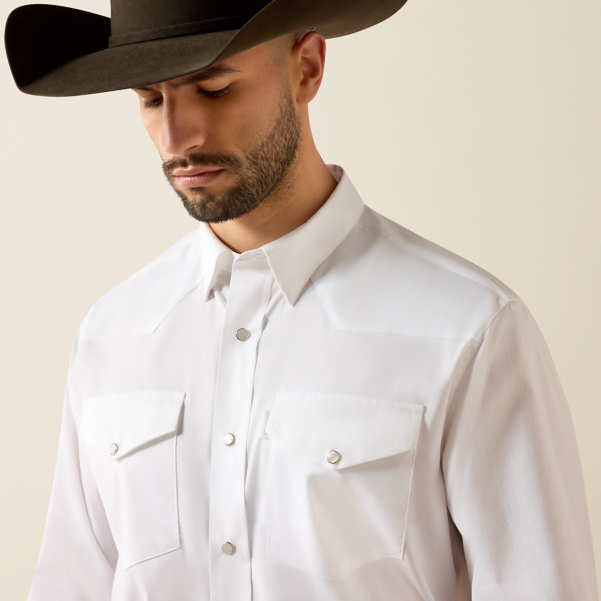 A man wearing a white western-style button-down shirt with two chest pockets and a black cowboy hat, photographed against a plain light beige background.