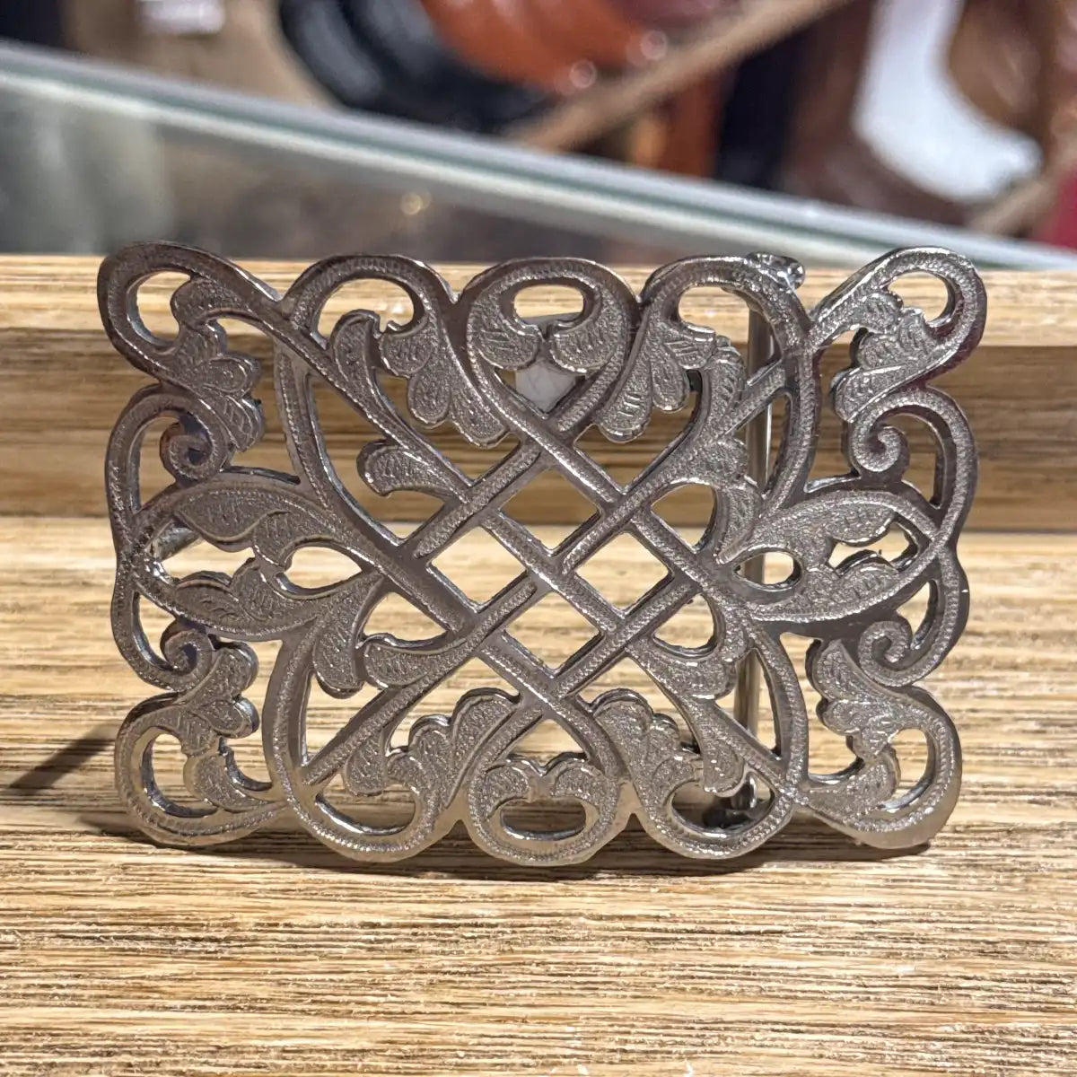 In-store counter display view of the handcrafted stainless steel cutout lattice cross western belt buckle on a wooden surface.