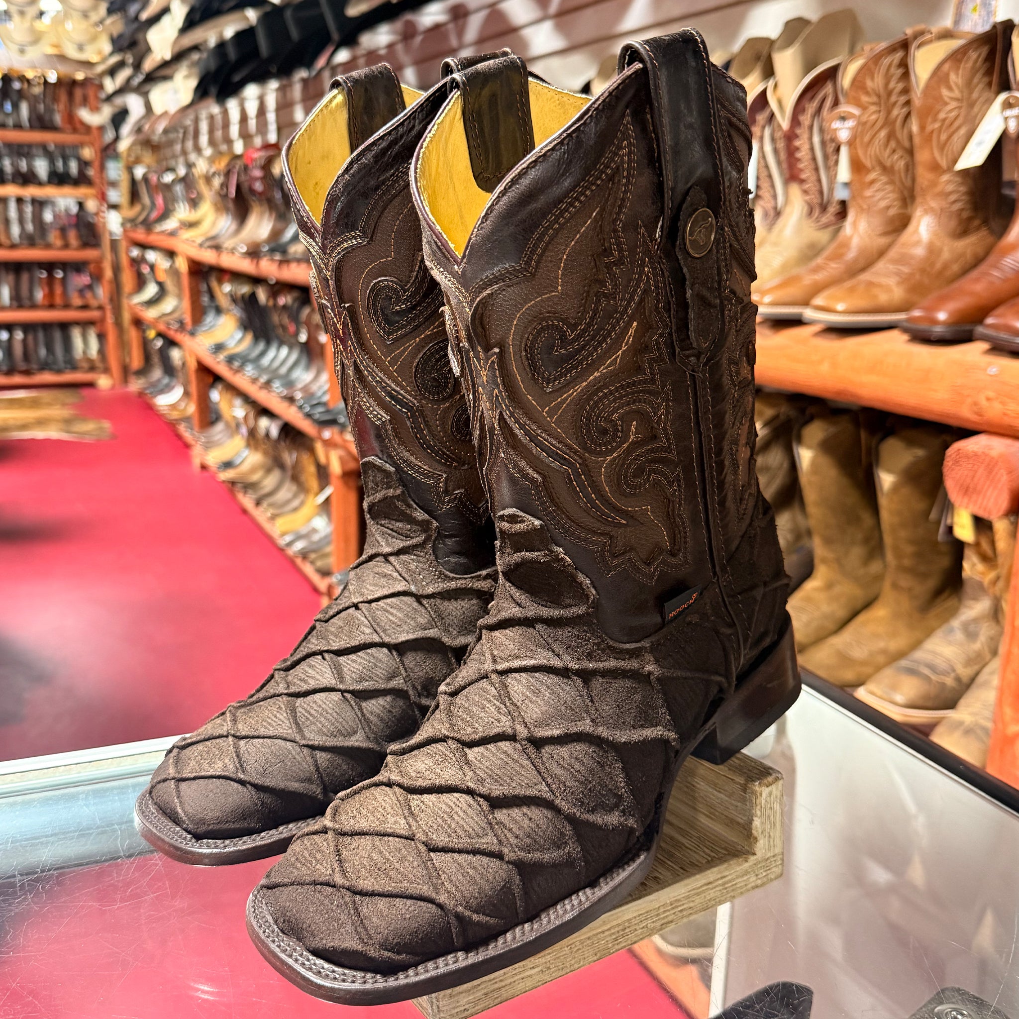 A dark brown square toe Pirarucu fish print cowboy boot with intricate hand-tooled western embroidery on the shaft, shown against a boot store background.