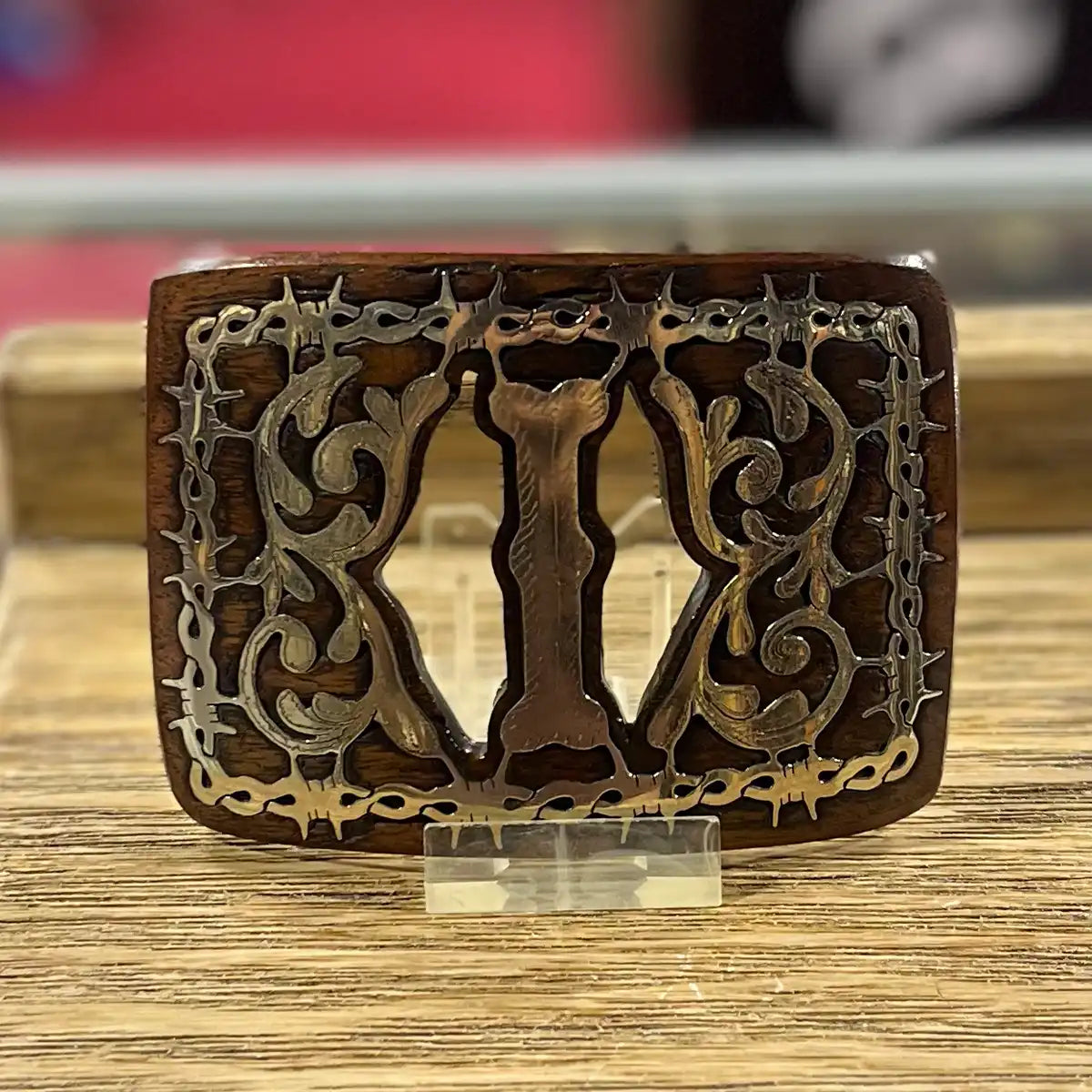 A rectangular wooden belt western buckle with ornate filigree cutouts and a central I, displayed on a small clear stand on a wooden surface.