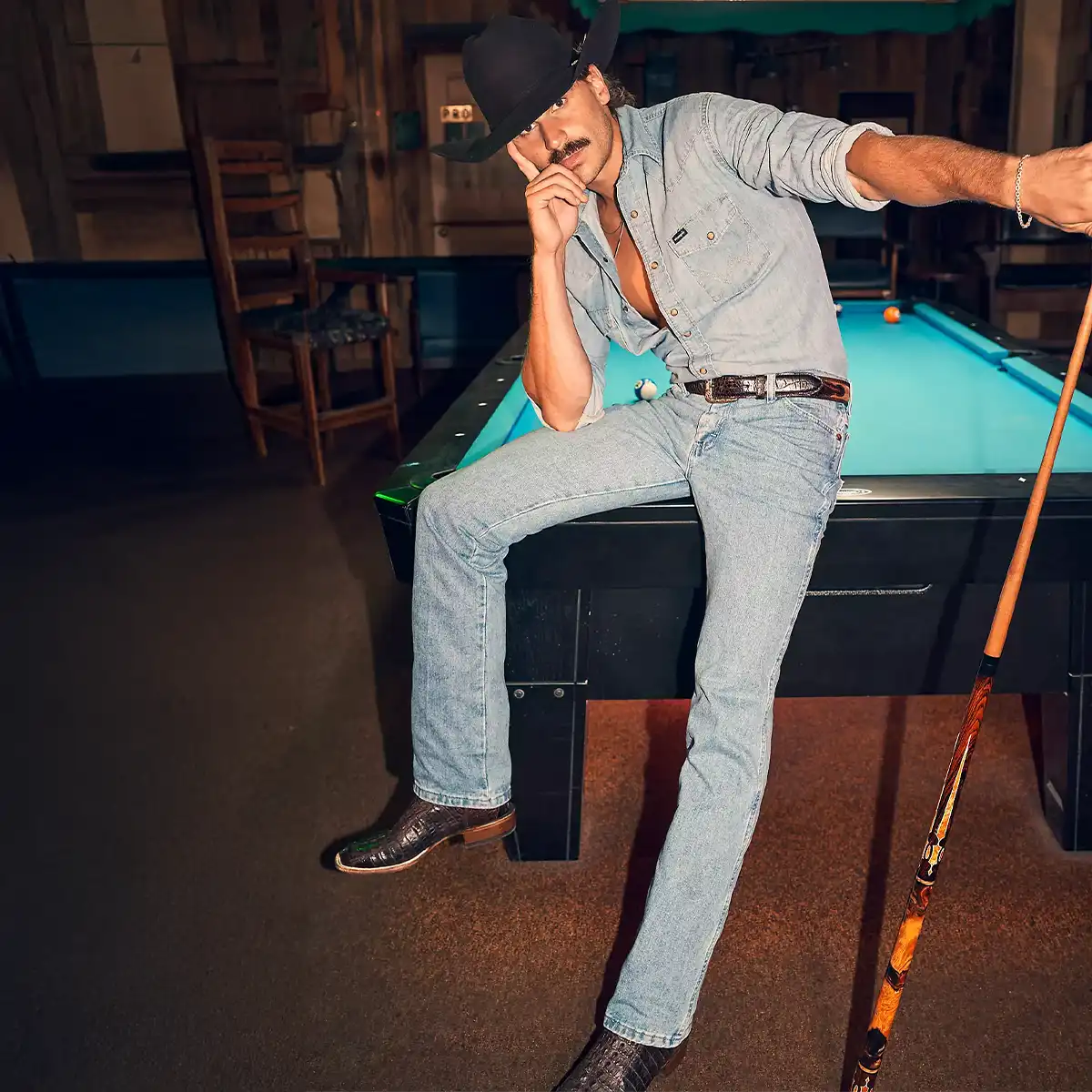 A man wearing a black cowboy hat, a light blue denim shirt, light blue Wrangler jeans, and black cowboy boots sits on a pool table in a dimly lit bar or lounge setting.