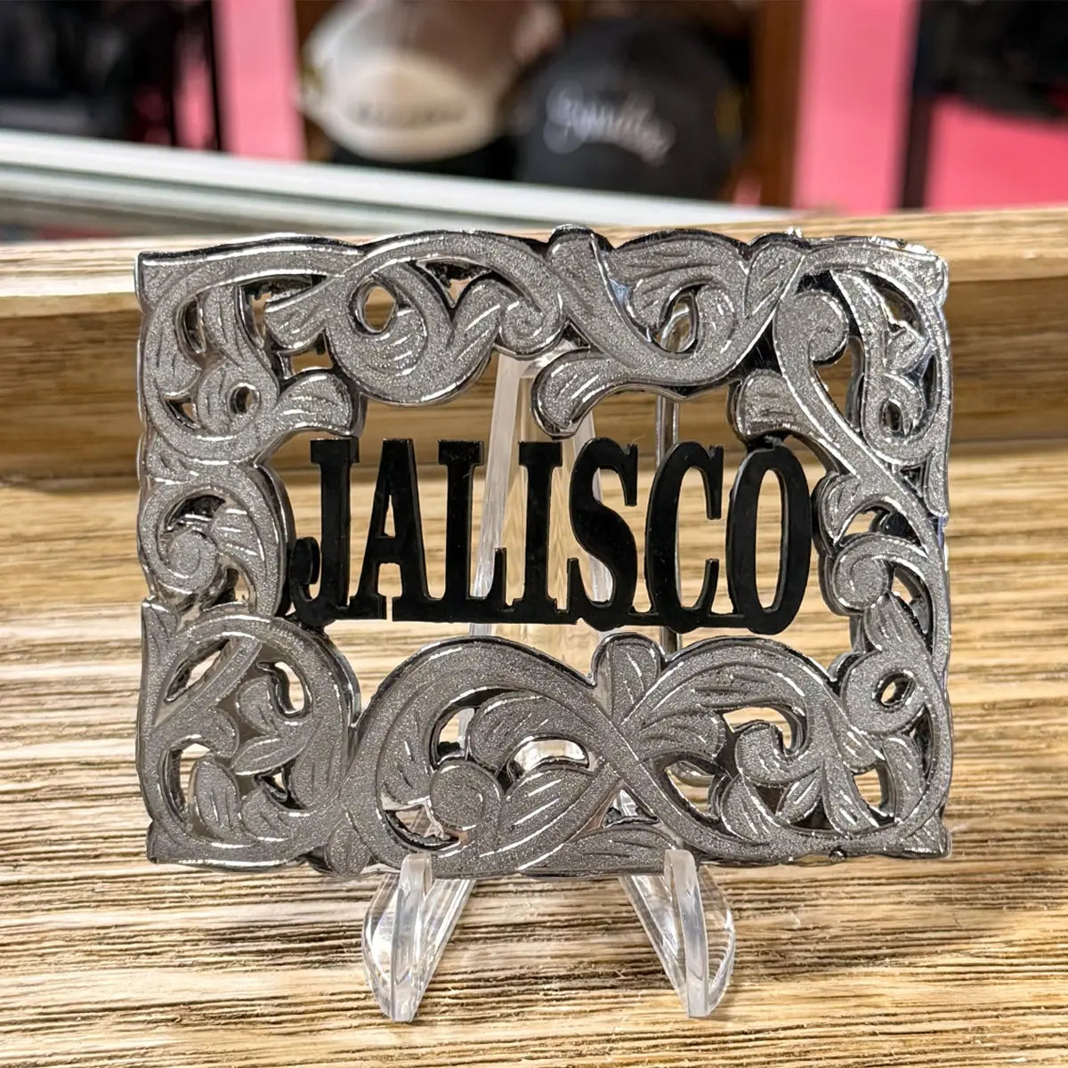Close-up display view of the Jalisco silver and black Western belt buckle held on an acrylic stand over a wooden counter.