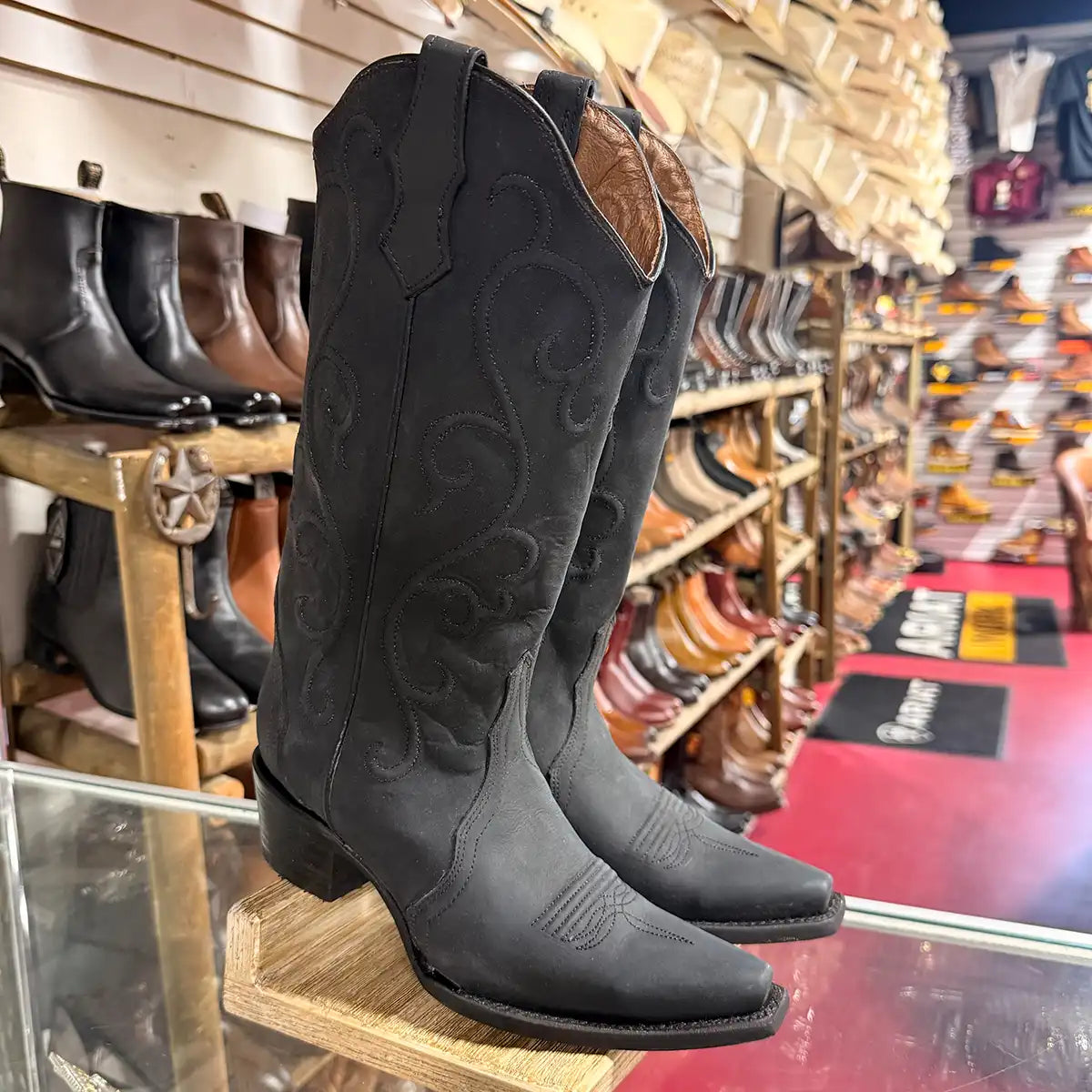 A pair of Corral women's black western cowboy boots with decorative stitching, displayed on a wooden stand inside a western store.