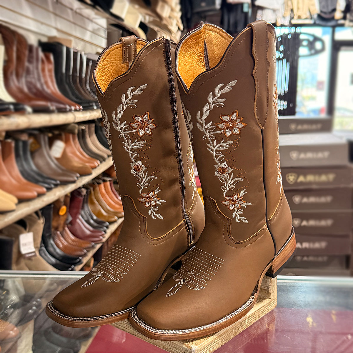 A pair of women's brown leather cowboy boots with floral embroidery on the shafts, displayed on a wooden riser in a retail western store setting with shelves of other boots in the background.