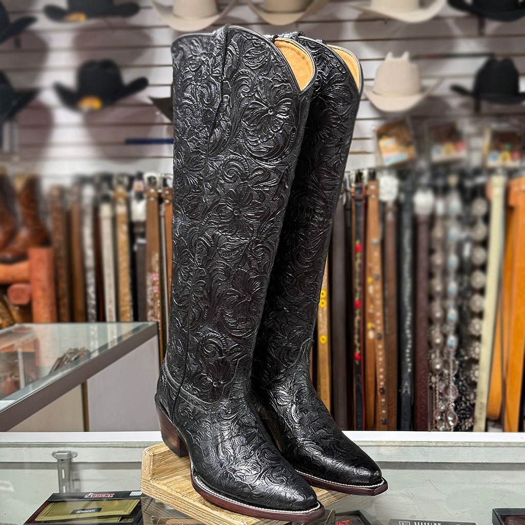 Retail display view of the Lirac black 17-inch tooled boots sitting on a glass counter inside a Western boot store surrounded by leather belts.