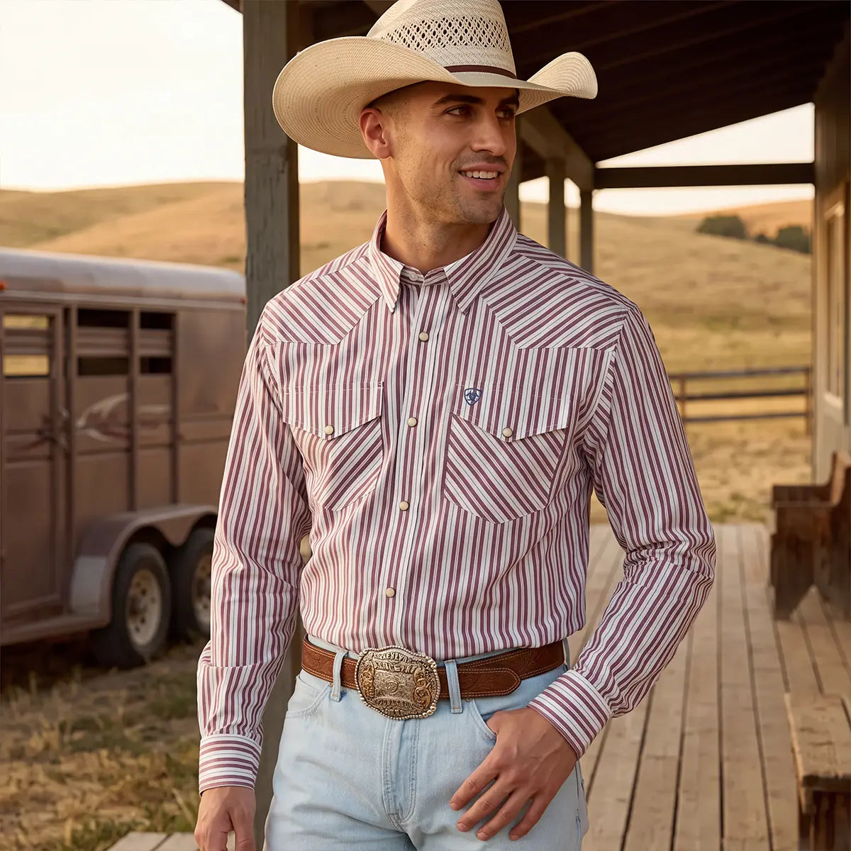 A man wearing a long-sleeve western shirt with red and white vertical stripes, a cowboy hat, and light blue jeans, standing on a wooden porch with a horse trailer in the background.