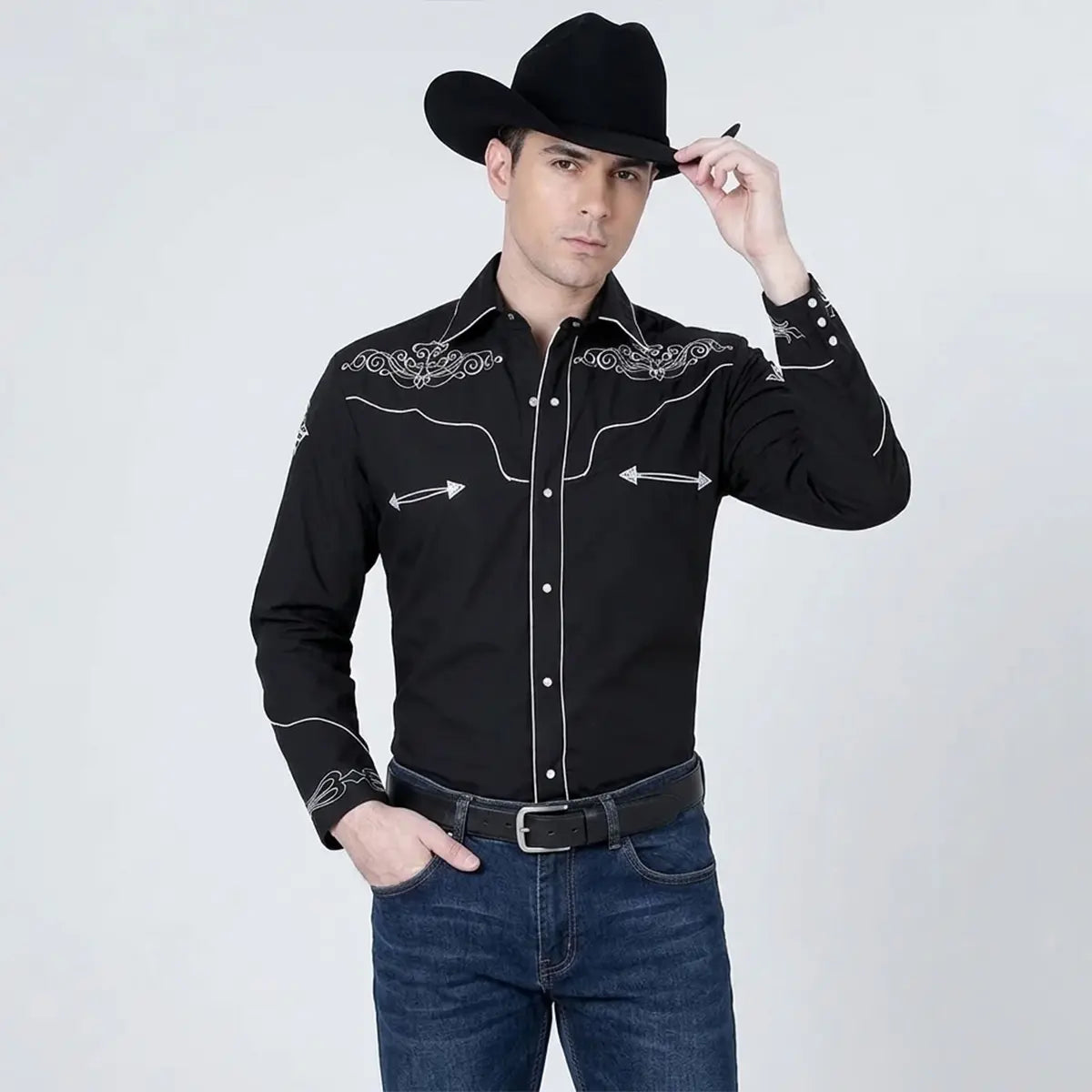 Front view of a male model wearing a men's long-sleeve black Western snap shirt featuring white scroll embroidery on the yokes, white piping on the placket, smiley arrow accents, black cowboy hat, black leather belt, and blue jeans.