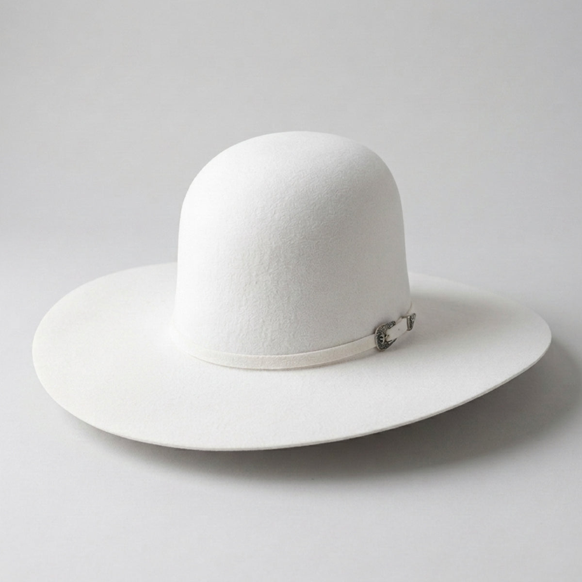 A Cuernos Chuecos white open crown felt cowboy hat with a matching white band and a small silver metal emblem, photographed on a light gray background.