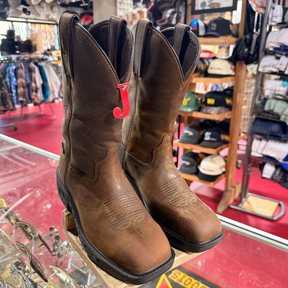 A pair of Justin brown leather cowboy boots displayed on a wooden stand inside a western store. The boots feature a rich brown color with subtle tonal stitching and a bright red J tag attached near the collar.