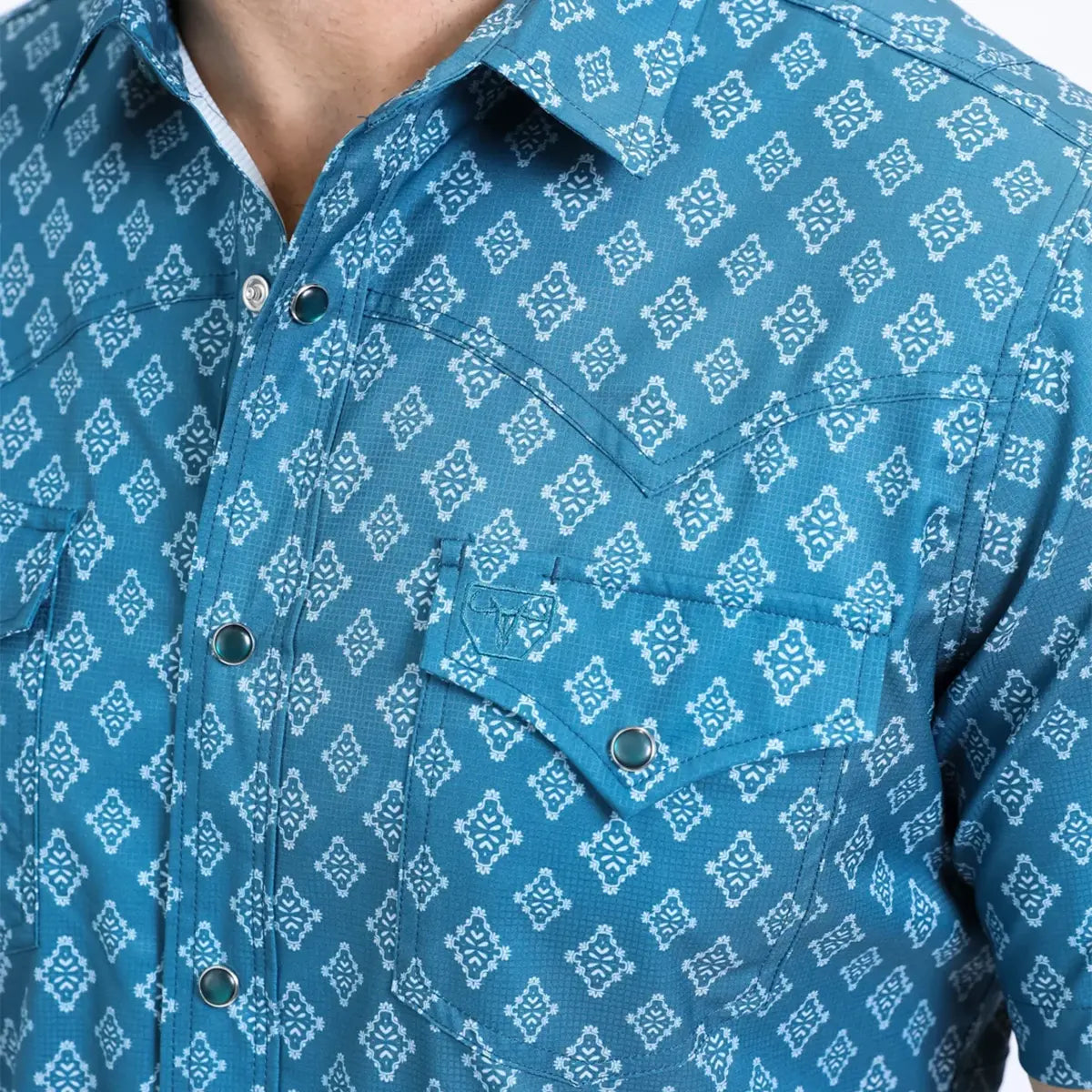 Close-up front chest view of the Platini blue Aztec performance shirt detailing the collar, amber snap button, Western yoke, and embroidered pocket logo.