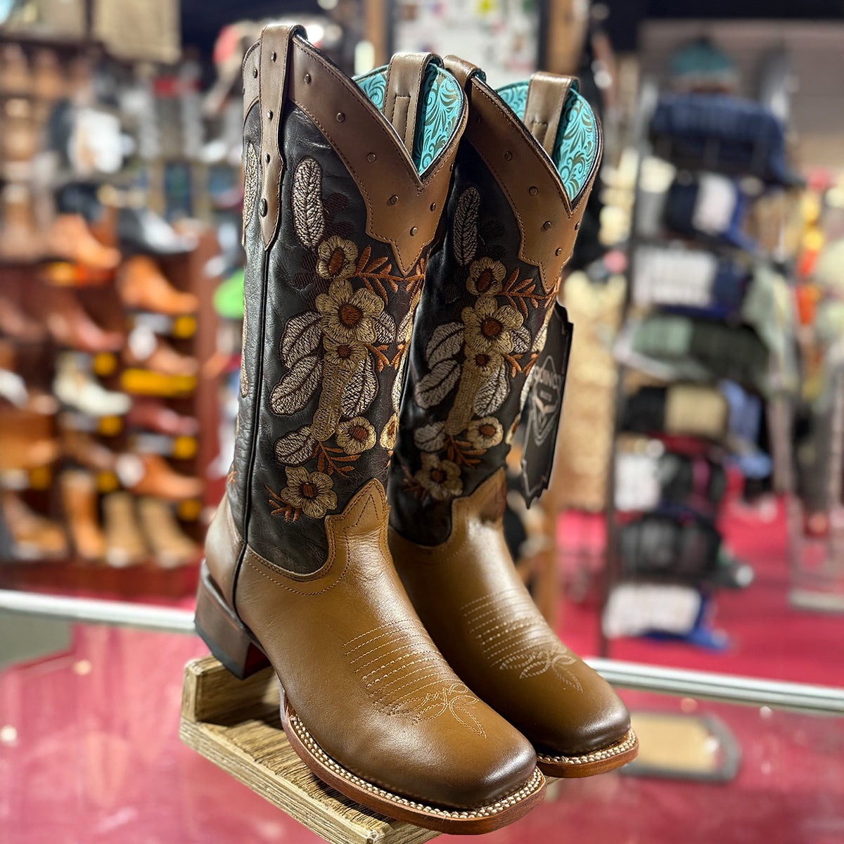 Pair of Quincy brown and black cowgirl square toe boots with floral patterns on a display stand in a western store.