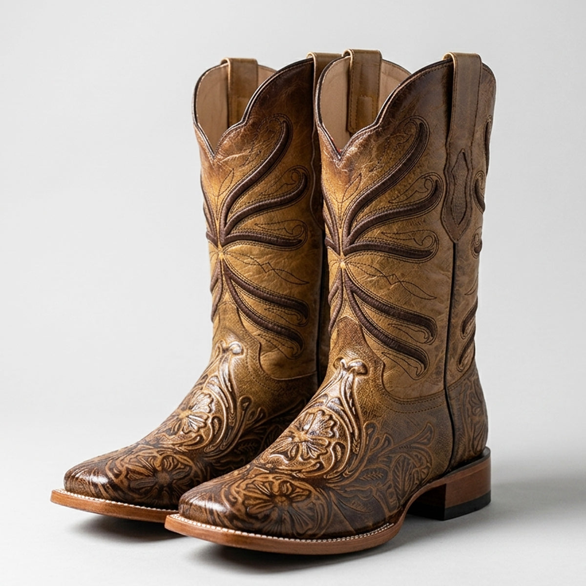 Pair of men's brown tooled square toe western boots with intricate designs on a light gray background.