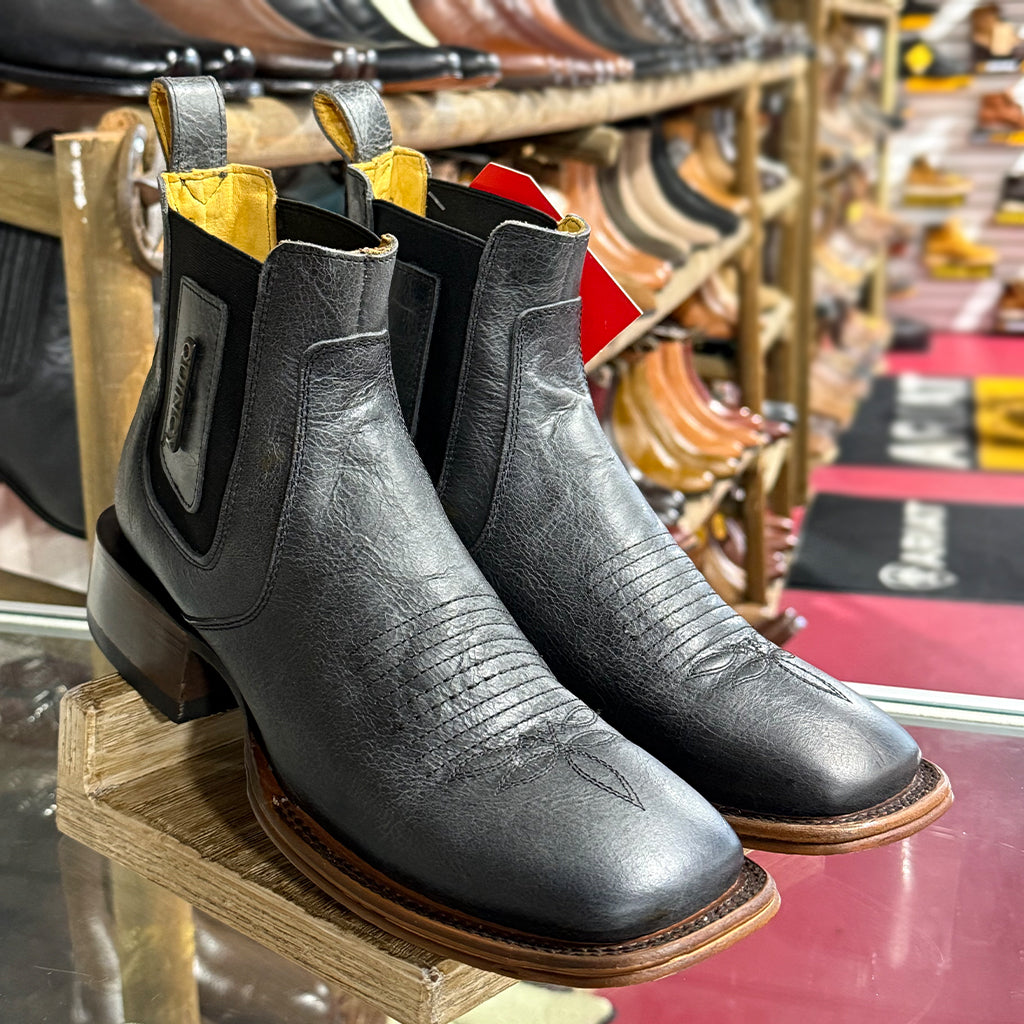 A pair of Quincy men's black leather cowboy Chelsea boots with a square toe and stacked block heel, displayed on a wooden stand in a western store.