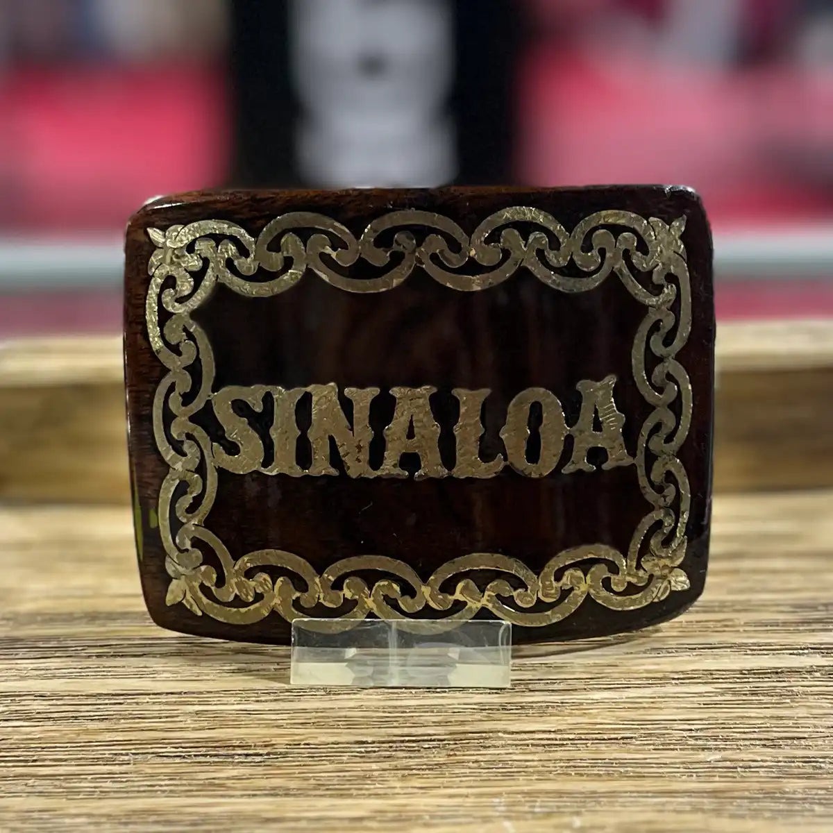 Front view of the handcrafted mesquite wood belt buckle with gold-tone metal SINALOA lettering display on a acrylic stand.