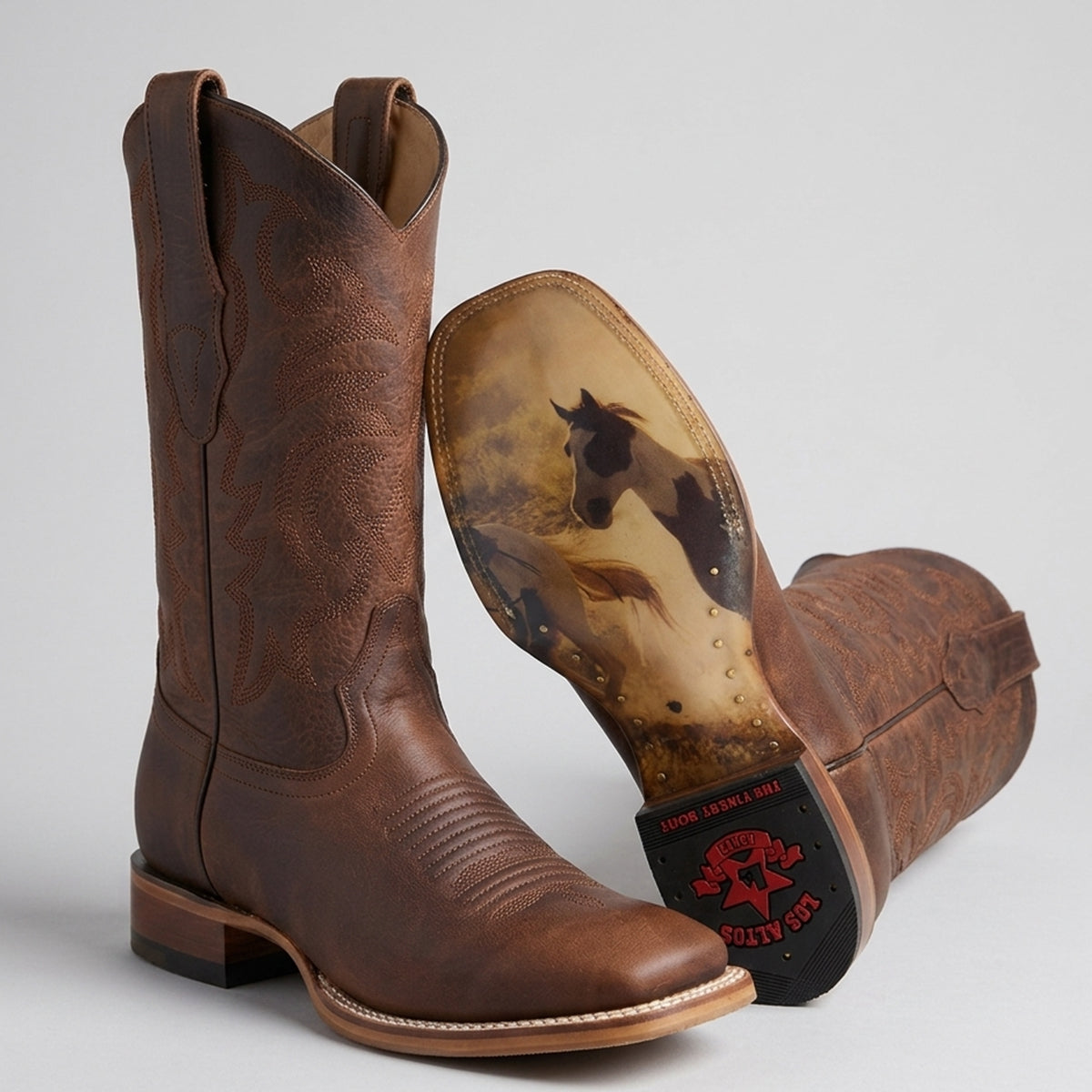 A pair of brown leather cowboy boots shown on a light gray studio background, with one boot standing upright and the other lying on its side to display the printed sole featuring a horse illustration.