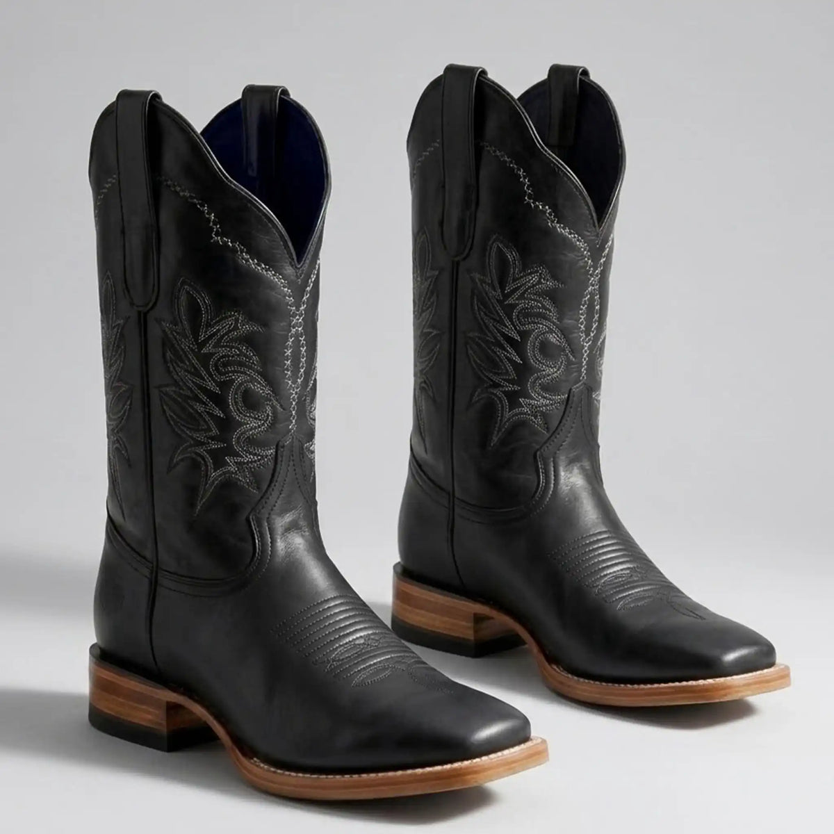 A pair of black leather cowboy boots with western embroidery, square toes, and stacked brown leather heels, shown on a light gray background.