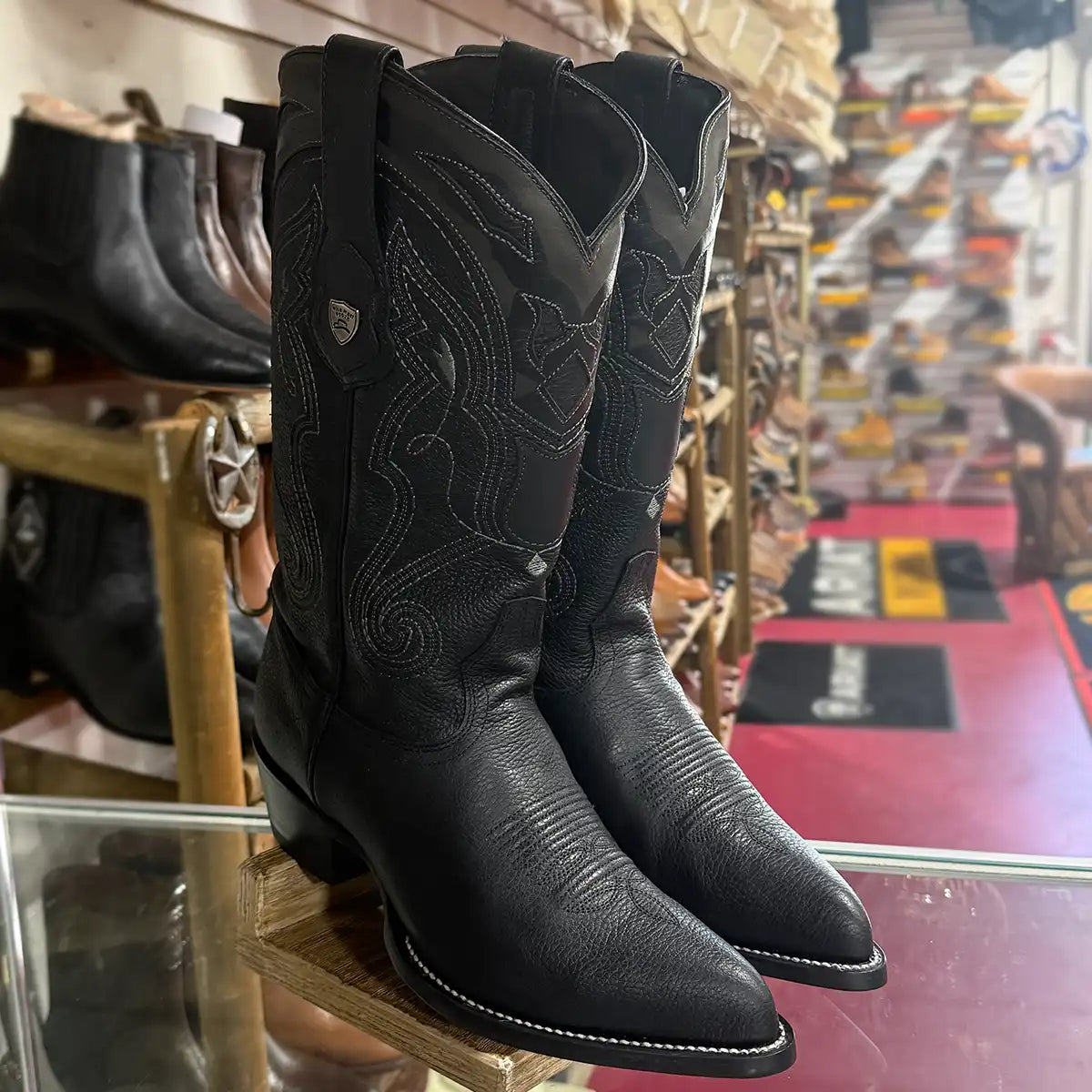A pair of black leather western boots with decorative stitching and a pointed toe, displayed on a glass counter inside a boot shop.