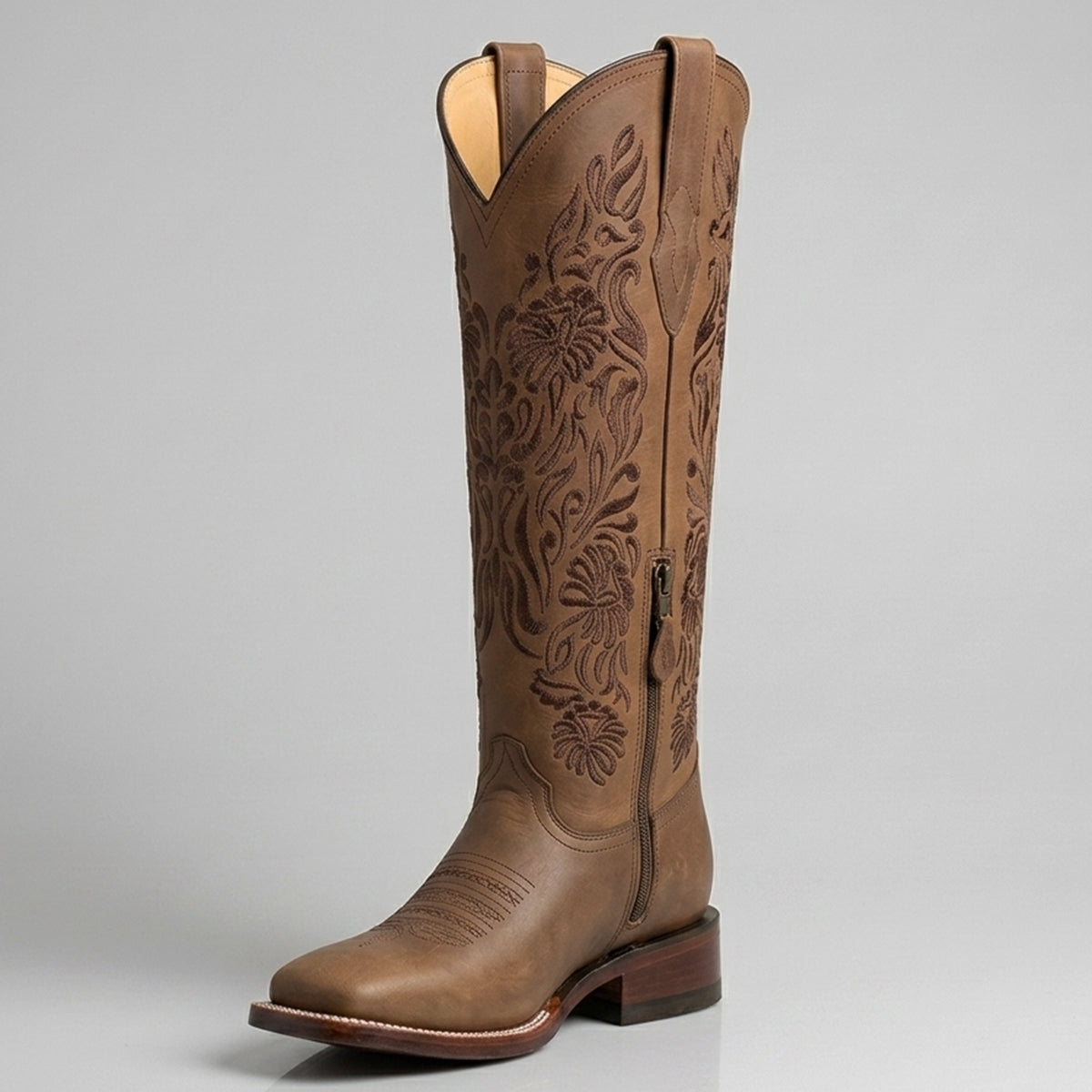 A brown leather women's cowboy boot with floral tooling on the shaft, shown from a three-quarter angle against a neutral gray background.