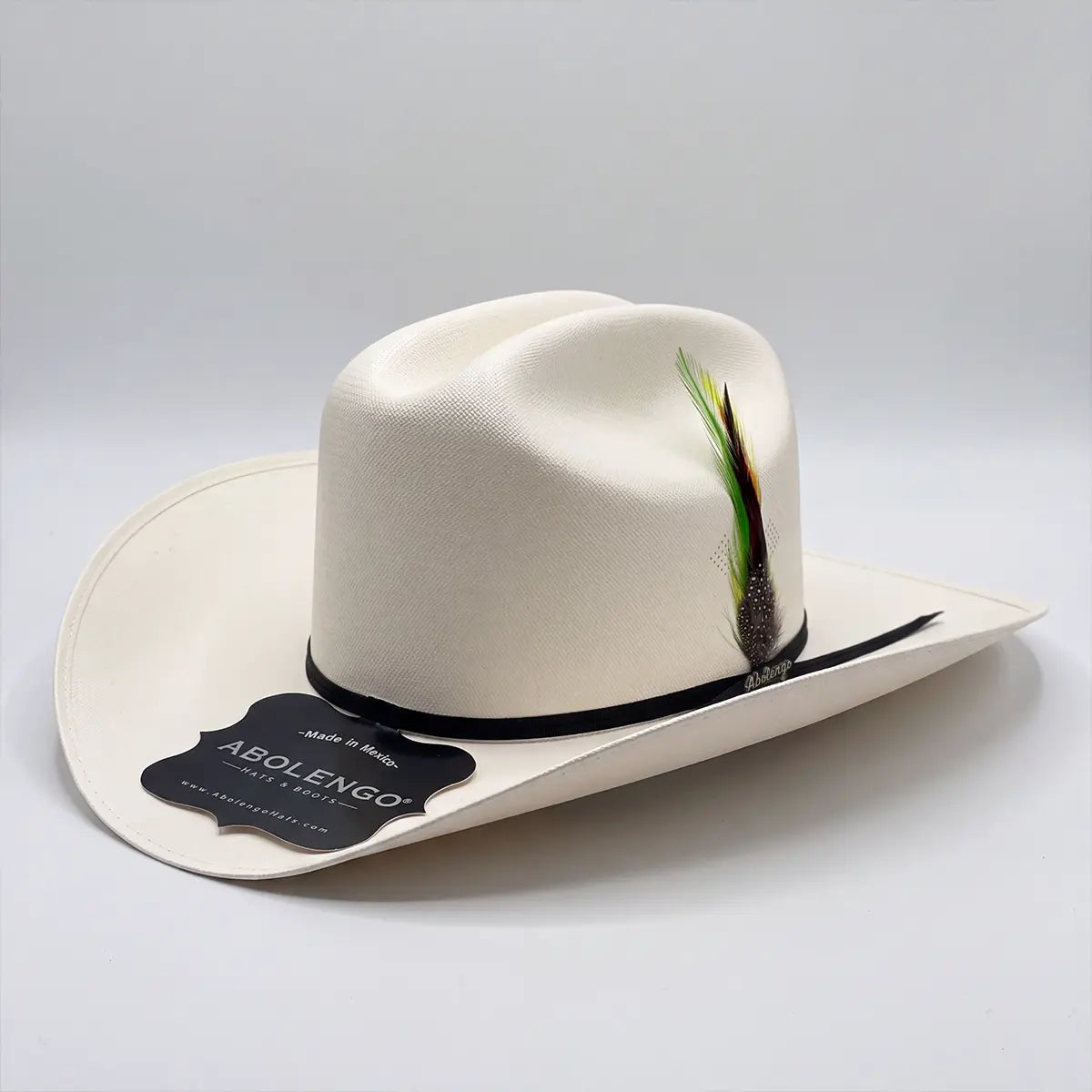 Three-quarter studio view of the Abolengo El Clásico 100X Sinaloa straw cowboy hat on a white background displaying the Cattleman crown, black ribbon band, and colorful feather accent.