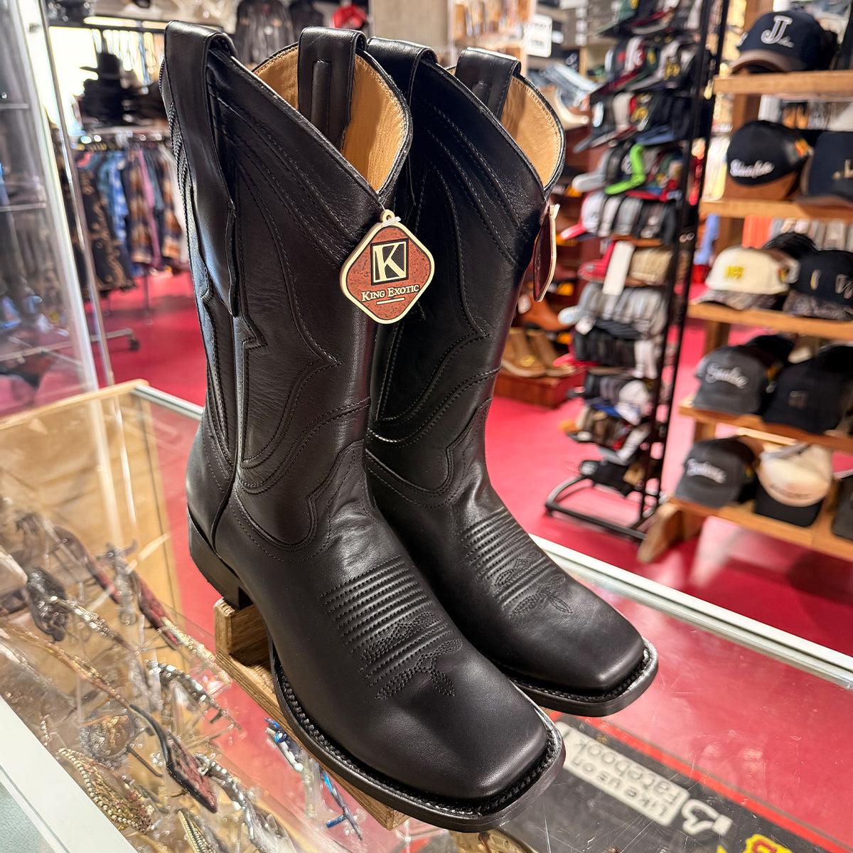 A pair of black leather cowboy boots displayed on a glass counter inside a western store. The boots feature decorative stitching, a square toe, and a hanging tag that reads 'King Exotic'.