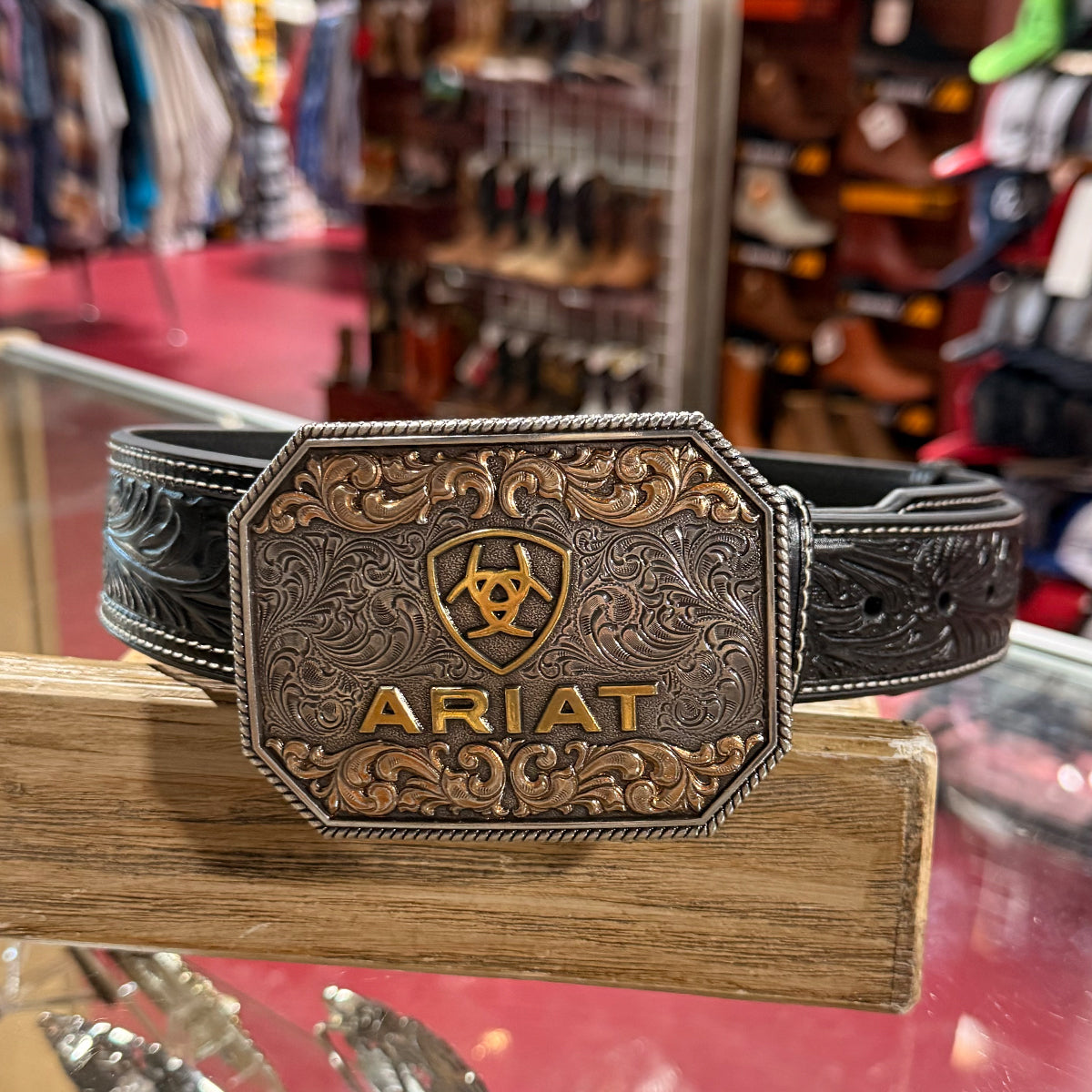 An Ariat western belt with an ornate engraved buckle resting on a wooden display stand inside a store.
