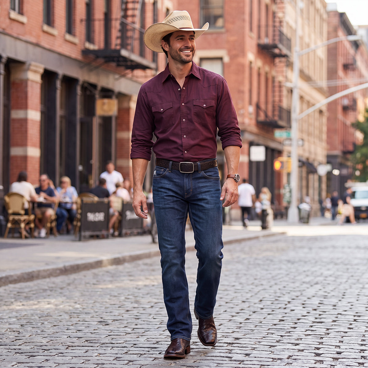 A smiling man wearing a long-sleeve burgundy western snap shirt, dark blue jeans, a dark brown leather belt, black cherry full quill ostrich cowboy boots, and a straw cowboy hat walking down a cobblestone street.