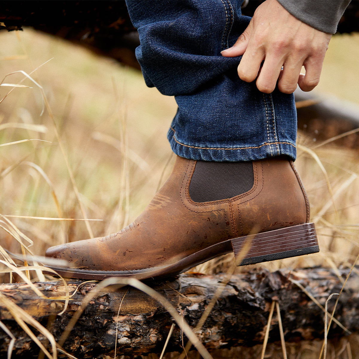 A close-up of a person wearing a brown leather Chelsea Ariat boot with dark blue jeans, standing on a log in an outdoor grassy setting.
