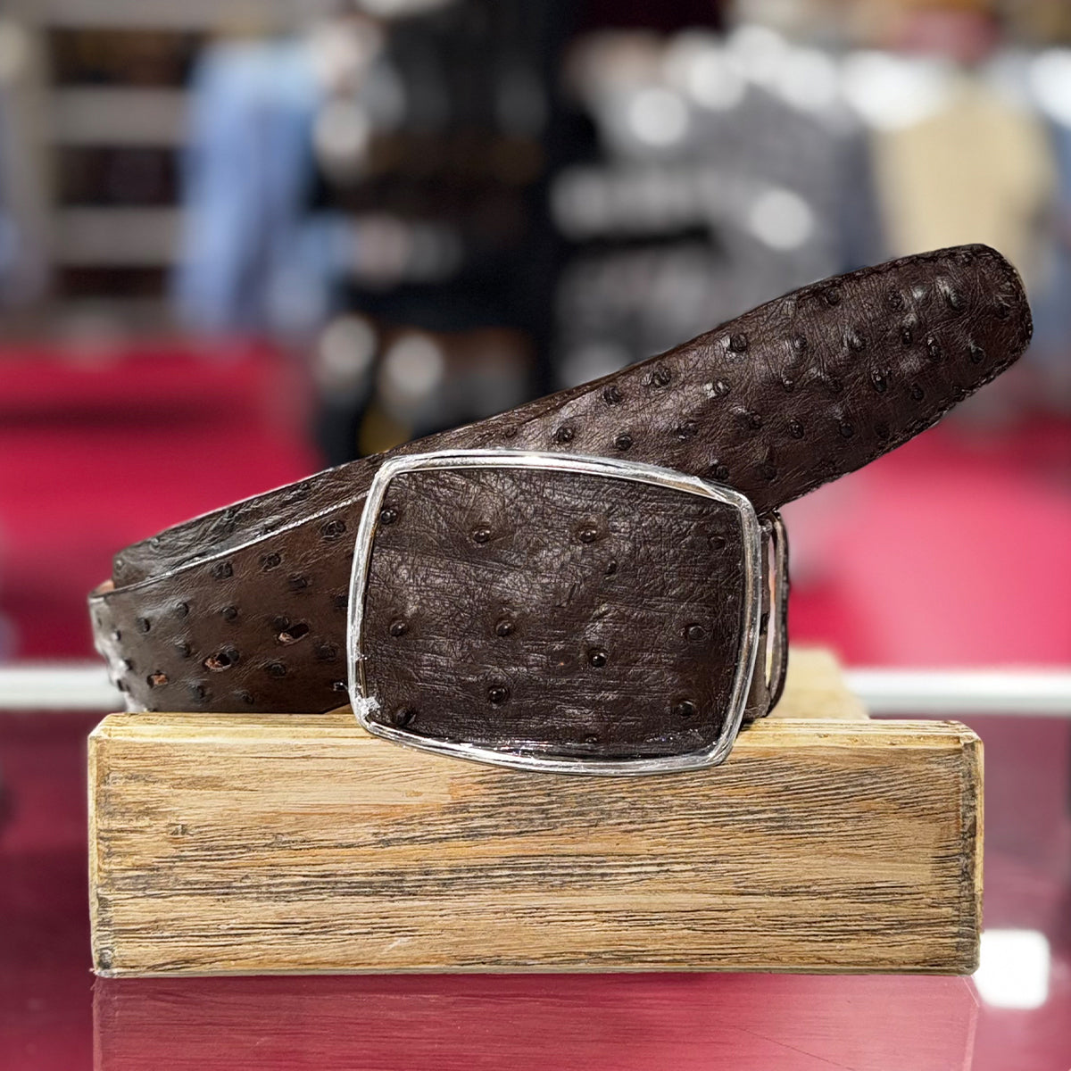 Los Altos brown full quill ostrich leather belt with a silver buckle on a wooden block against a blurred background.