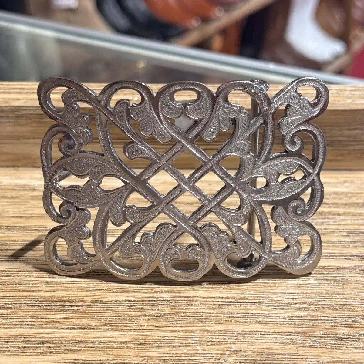 An ornate silver-toned metal belt buckle with a detailed filigree knot design, resting on a wooden surface.