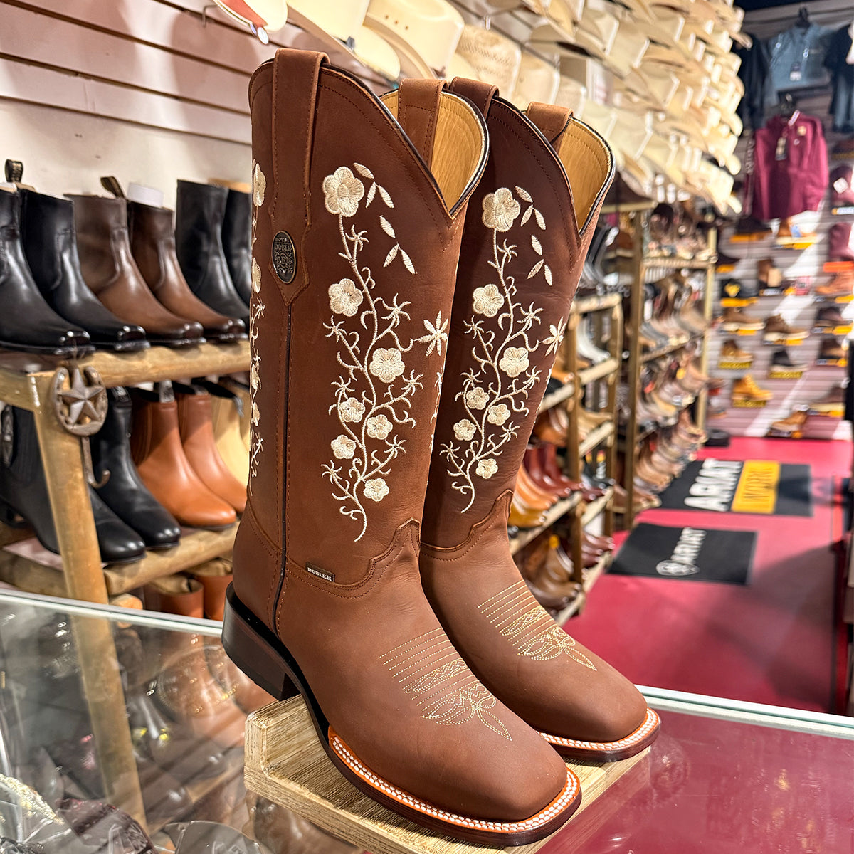 A pair of women's brown leather square toe cowgirl boots with white floral embroidery, displayed on a wooden stand inside a western store.