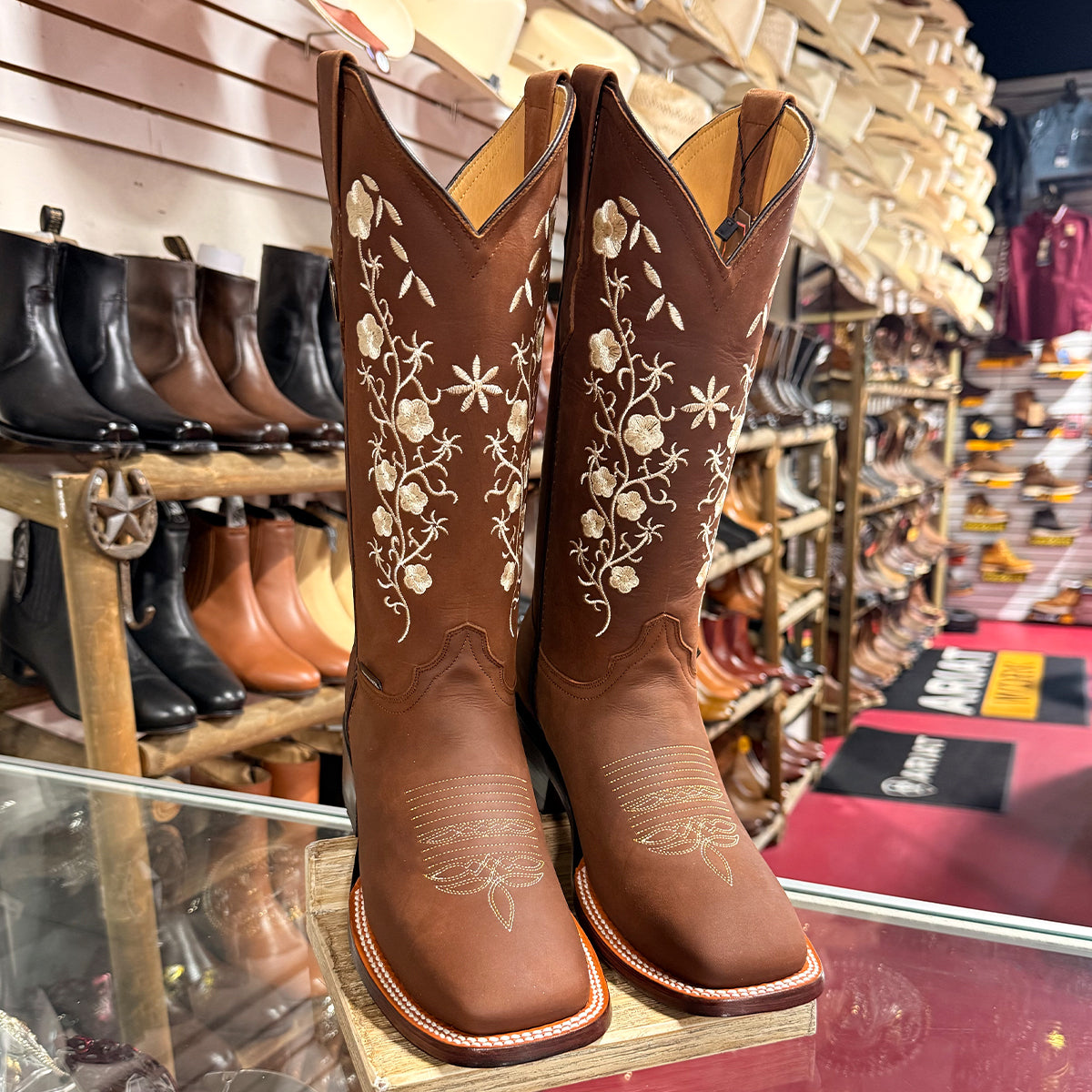 A pair of women's brown leather cowboy boots with white floral embroidery on the shafts, displayed on a wooden stand inside a western store.