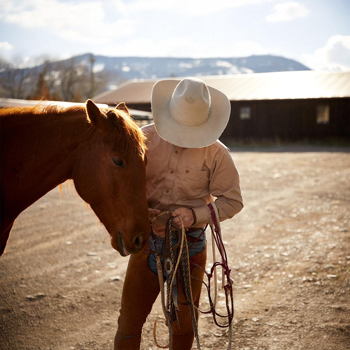 A cowboy in a light cowboy hat, Ariat tan button-down shirt, and chaps standing outdoors next to a chestnut horse while adjusting a leather lead rope.