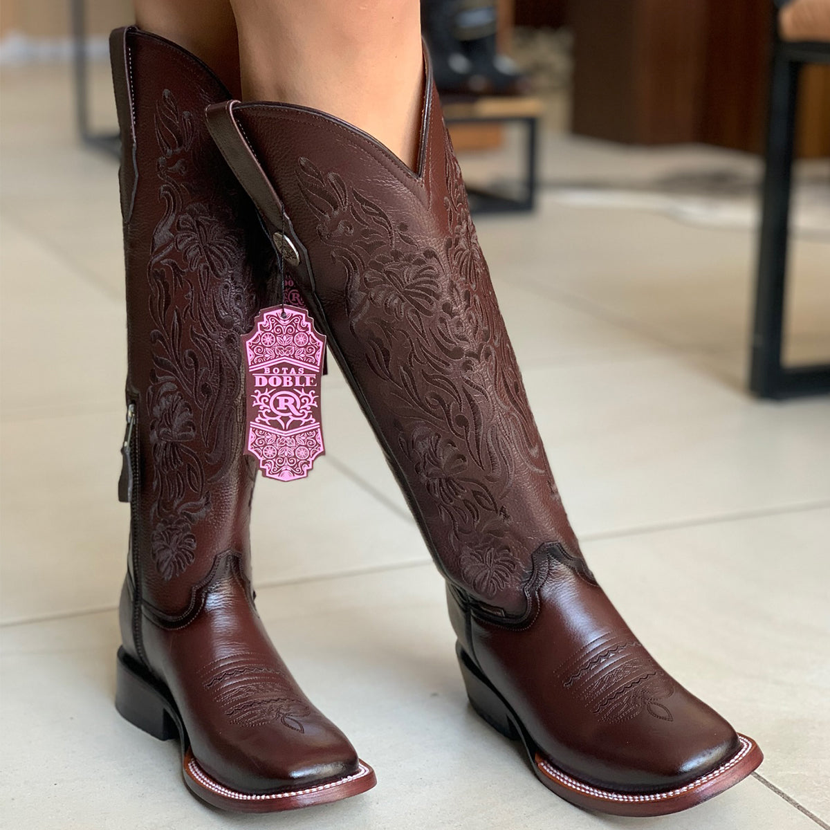 A pair of dark brown leather women's square toe cowboy boots with floral embroidered design shown on a model's feet. A 'Botas Doble R' brand tag hangs from the shaft of the left boot.
