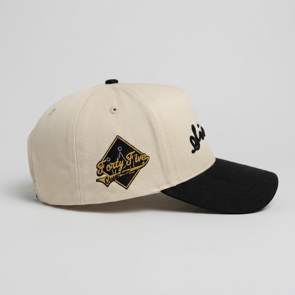 A structured baseball cap in cream and black, featuring a black curved brim, a black button on top, a black embroidered script logo on the side, and a black diamond patch with gold and black text on the front.