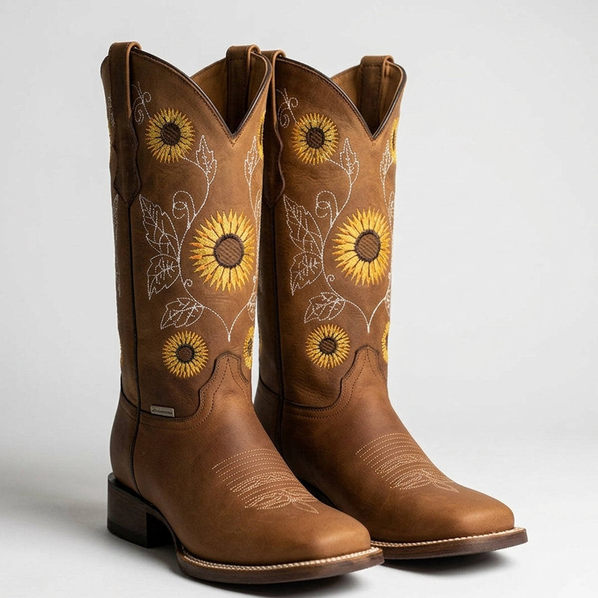 A pair of women's brown leather cowboy boots with embroidered yellow sunflowers and white floral details on the shafts, shown against a light gray background.