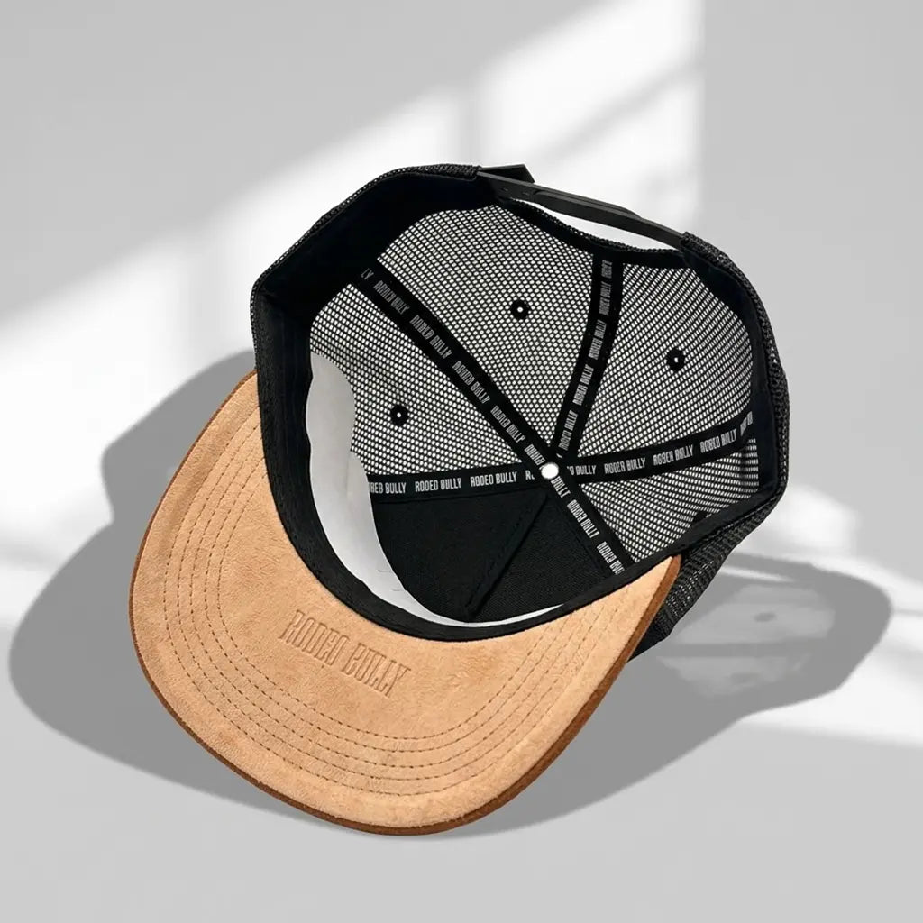 Underside studio view of the Rodeo Bully black trucker cap displaying the heat-stamped "RODEO BULLY" logo on the brown suede brim, branded seam tape, inner sweatband, and adjustable snapback closure.