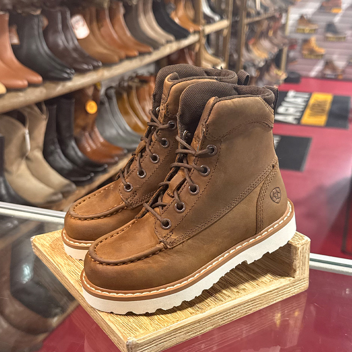 Kids Ariat brown leather boots on a wooden block with shelves of shoes in the background.