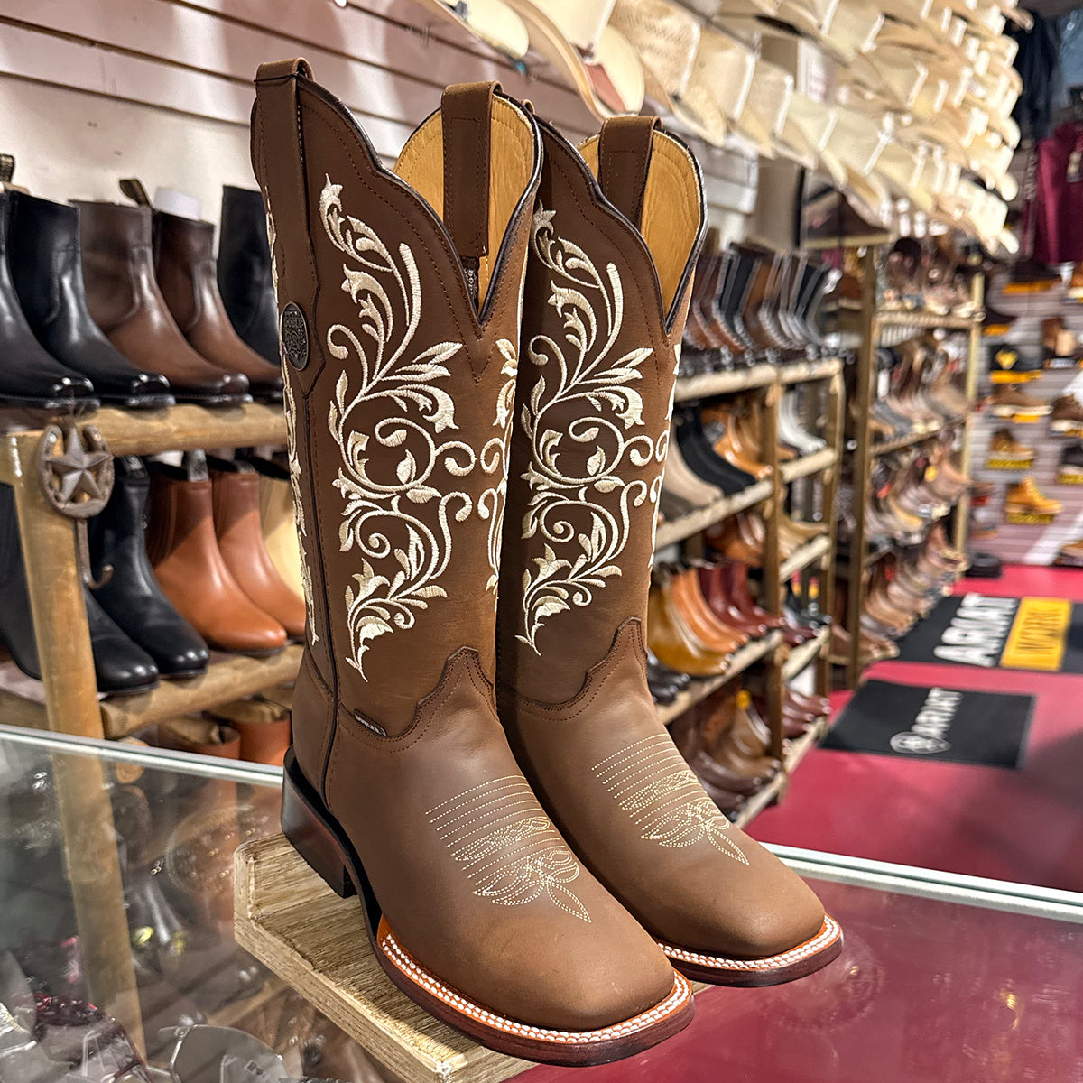A pair of women's Doble R brown leather cowboy boots with white floral embroidery, displayed on a wooden stand inside a shoe store.