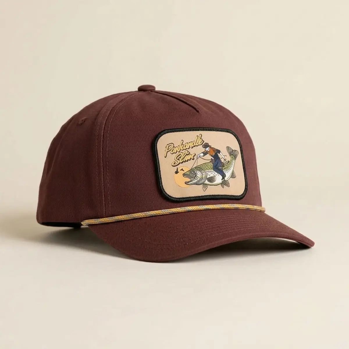 Front profile view of the Panhandle Slim maroon 5-panel cap displaying the cowboy riding a fish graphic patch and yellow braided rope cord.