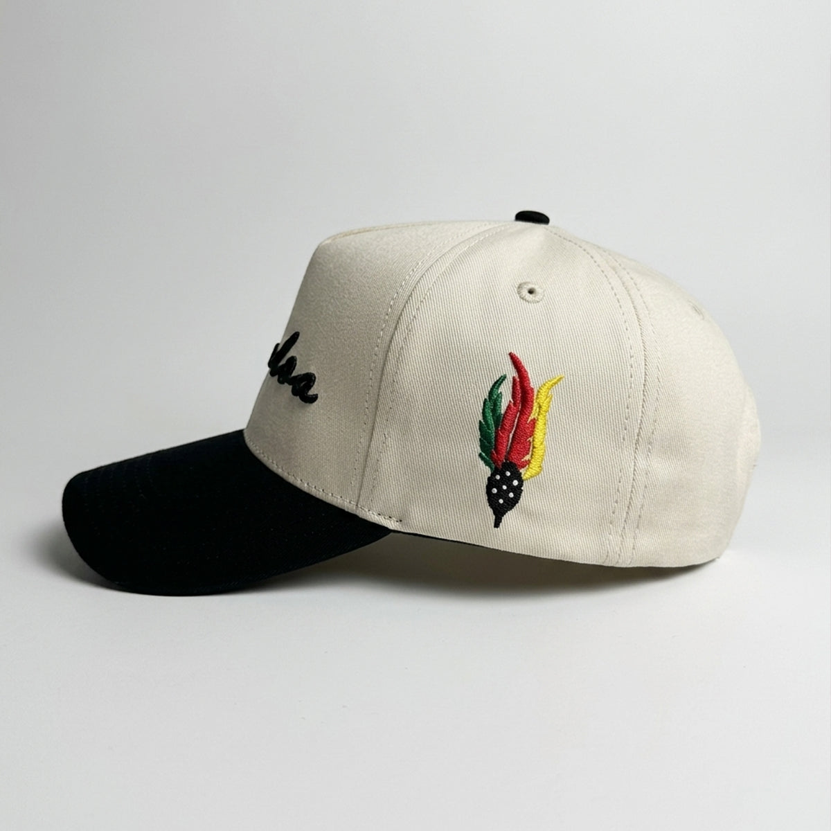 An off-white baseball cap with a black visor, featuring an embroidered black script logo on the side and a colorful feather patch in red, yellow, green, and black on the front.